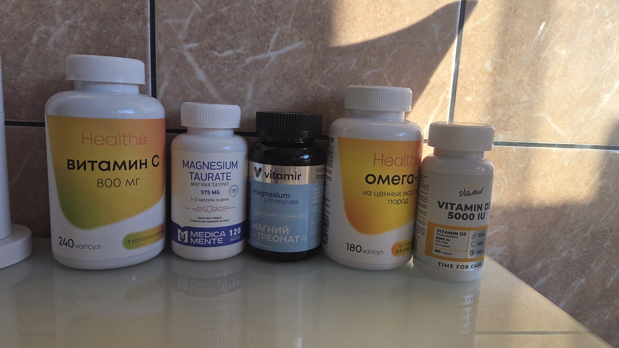 The image shows five bottles of supplements lined up on a glossy surface, likely a countertop or shelf. From left to right, the bottles contain: Healthis Vitamin C 800 mg, Medica Mente Magnesium Taurate 575 mg, Vitamir Magnesium L-Threonate, Healthis Omega, and Vita Meal Vitamin D3 5000 IU. The bottles are of varying sizes and shapes, with white or black caps. The background consists of a tiled wall. The image was likely taken indoors. There is no information in the photo regarding the city of Chaikovsky, Russia.