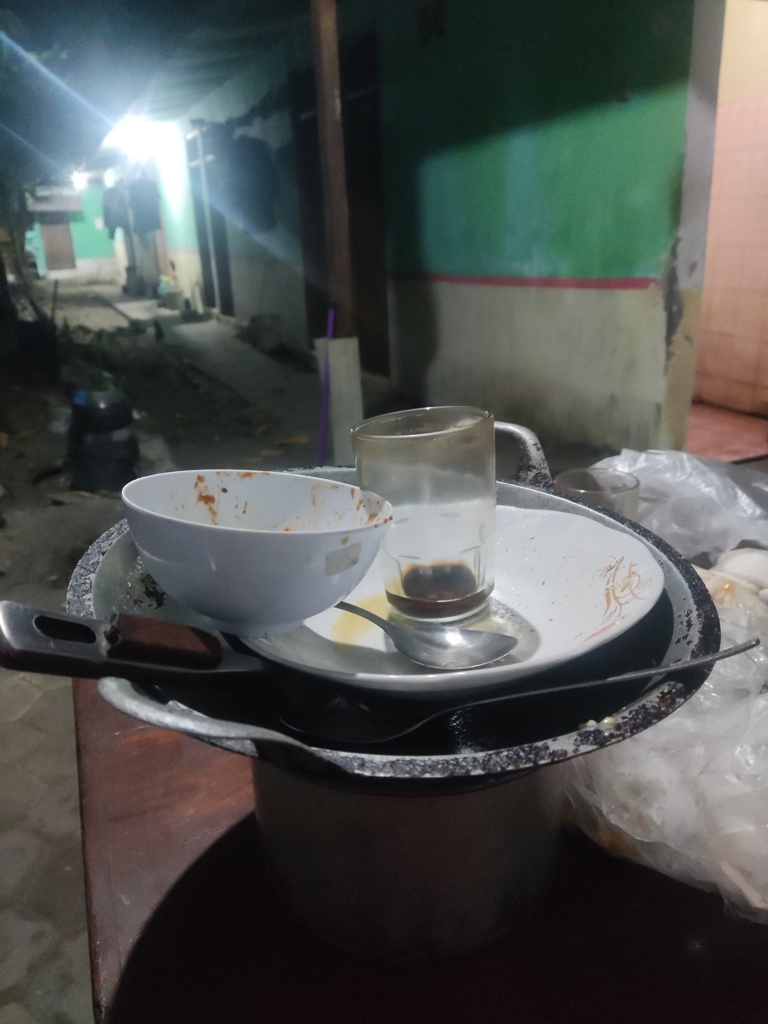 The image depicts a close-up view of dirty dishes stacked on a table, with a blurry outdoor scene in the background. The dishes include a white bowl with remnants of red sauce, a white plate with a metallic pattern, and a glass containing a small amount of dark liquid, likely coffee. A spoon rests on the plate, and a fork is partially visible on the left. The dishes are placed on a larger, dark-colored tray or pan with a patterned rim. In the background, a dimly lit outdoor area is visible, with a green and yellowish wall, some structures, and what appears to be a pathway. The lighting suggests it is either nighttime or an indoor setting with minimal illumination. The presence of plastic bags on the right side of the image indicates a potential outdoor setting or a storage area. The overall impression is that of a recently used eating area, with the dishes left uncleaned. The image is taken from a slightly elevated perspective, looking down at the dishes. The location is identified as Trihanggo, Indonesia.