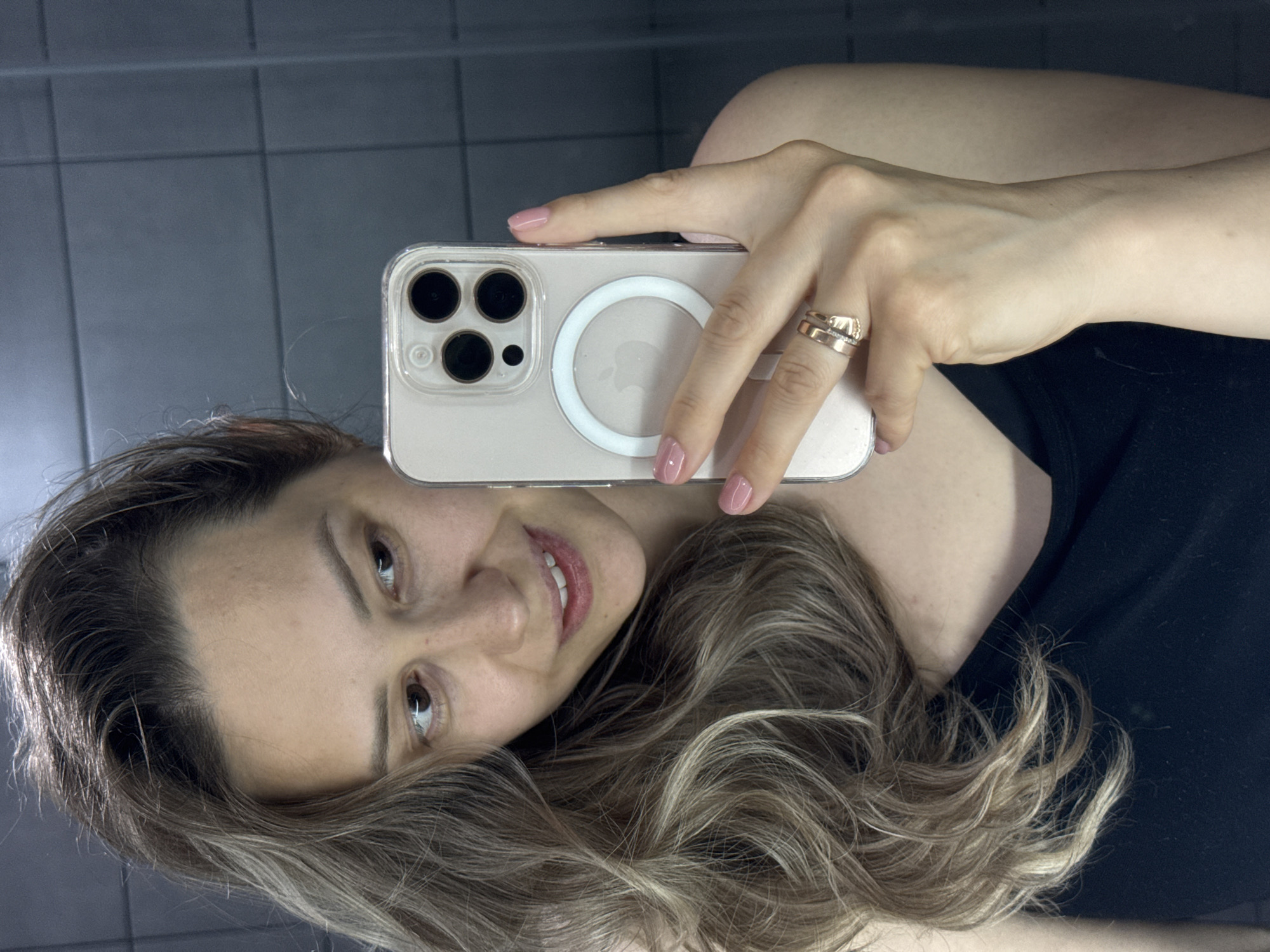 An indoor mirror selfie shows a woman holding a smartphone, reflecting her upper body and face. The woman has light brown, wavy hair with blonde highlights and a gentle smile, looking towards the camera. She has light-colored eyes and is wearing a black top or dress. Her fingernails are painted light pink, and she wears two gold-toned rings, one with what appears to be a diamond band, on her right ring finger.
She is holding a light-colored iPhone, likely a newer model given the triple camera setup, encased in a clear protective case. A white MagSafe ring is prominent on the back of the phone case, centered over the partially visible Apple logo. The background, visible around her head and behind the phone, consists of grey tiled walls, suggesting a bathroom or a similar utility space. The lighting is soft but adequate, highlighting her features. The setting is an interior space, with the general location context being Chaikovsky, Russia.