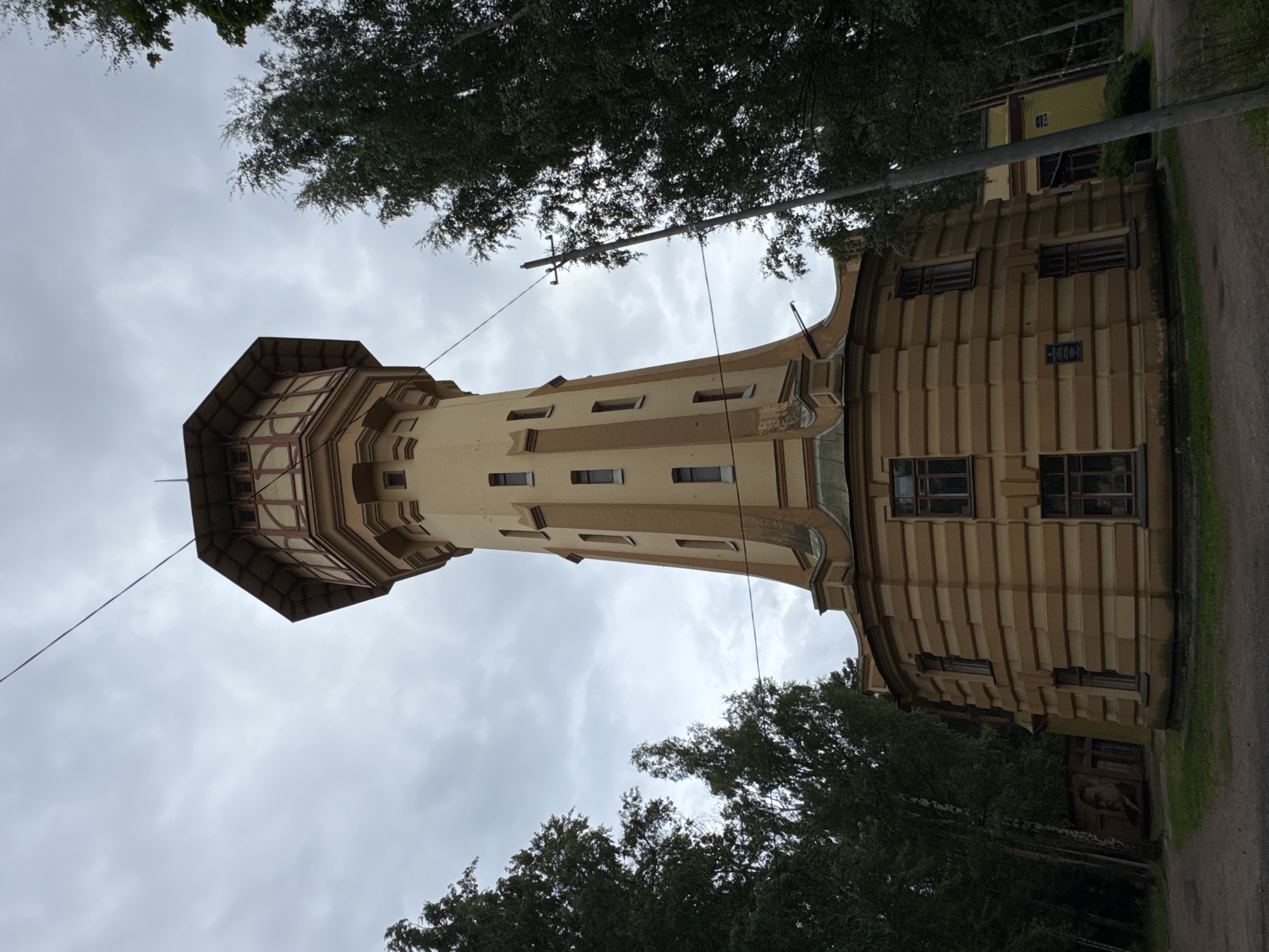 The image shows a historic water tower in a park-like setting, likely in Russia given the Cyrillic script on one of the buildings visible in the background. The water tower is painted in two shades of yellow and has an octagonal upper section with wooden detailing and windows. The base of the tower is wider and features a patterned facade with multiple windows. Surrounding the tower are numerous tall, green trees, and the sky is overcast and gray. In the lower left corner, a mural of a man's face is visible on a wall. The ground is covered with a dirt path and some grass. A telephone pole with wires runs diagonally across the image. The overall impression is one of a somewhat neglected but architecturally interesting historical landmark.