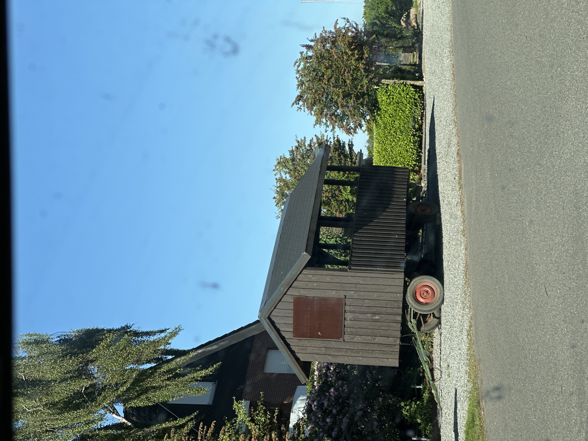The image shows a wooden structure with a pitched roof and a closed section with a square, reddish-brown panel in the middle, positioned beside a two-story building with visible windows and brickwork. The wooden structure is mounted on wheels, with a red wheel visible. To the right of the structure is a gravel area and a paved road. Various trees and bushes are present in the foreground and background. The location is Kutenholz, Germany.