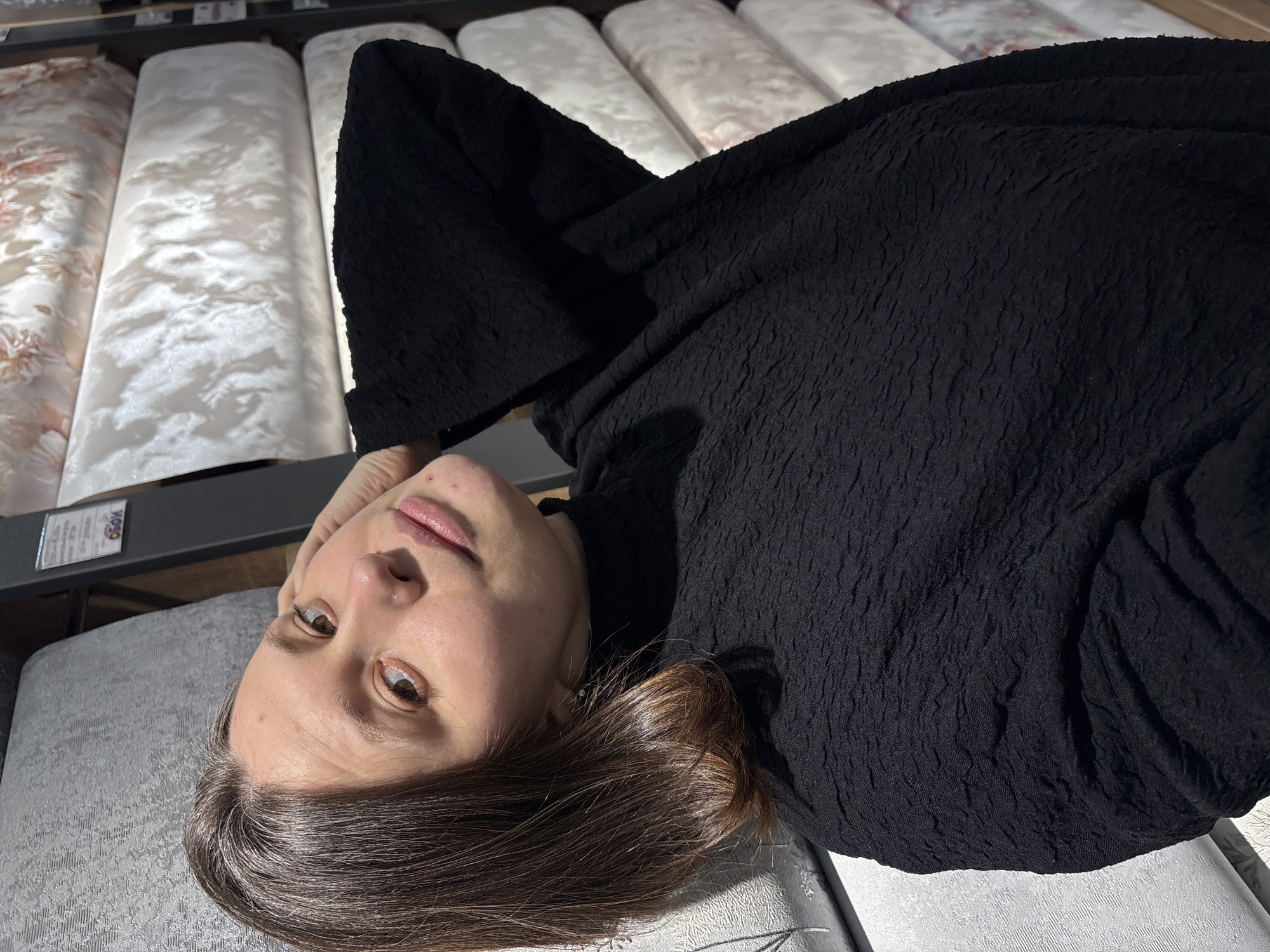 A woman lies on a gray quilted mattress, surrounded by a display of patterned mattresses at a store in Chaikovsky, Russia. She wears a black textured top. Her face is visible, and she is looking at the camera. The lighting is bright and appears to be a combination of natural and artificial light.