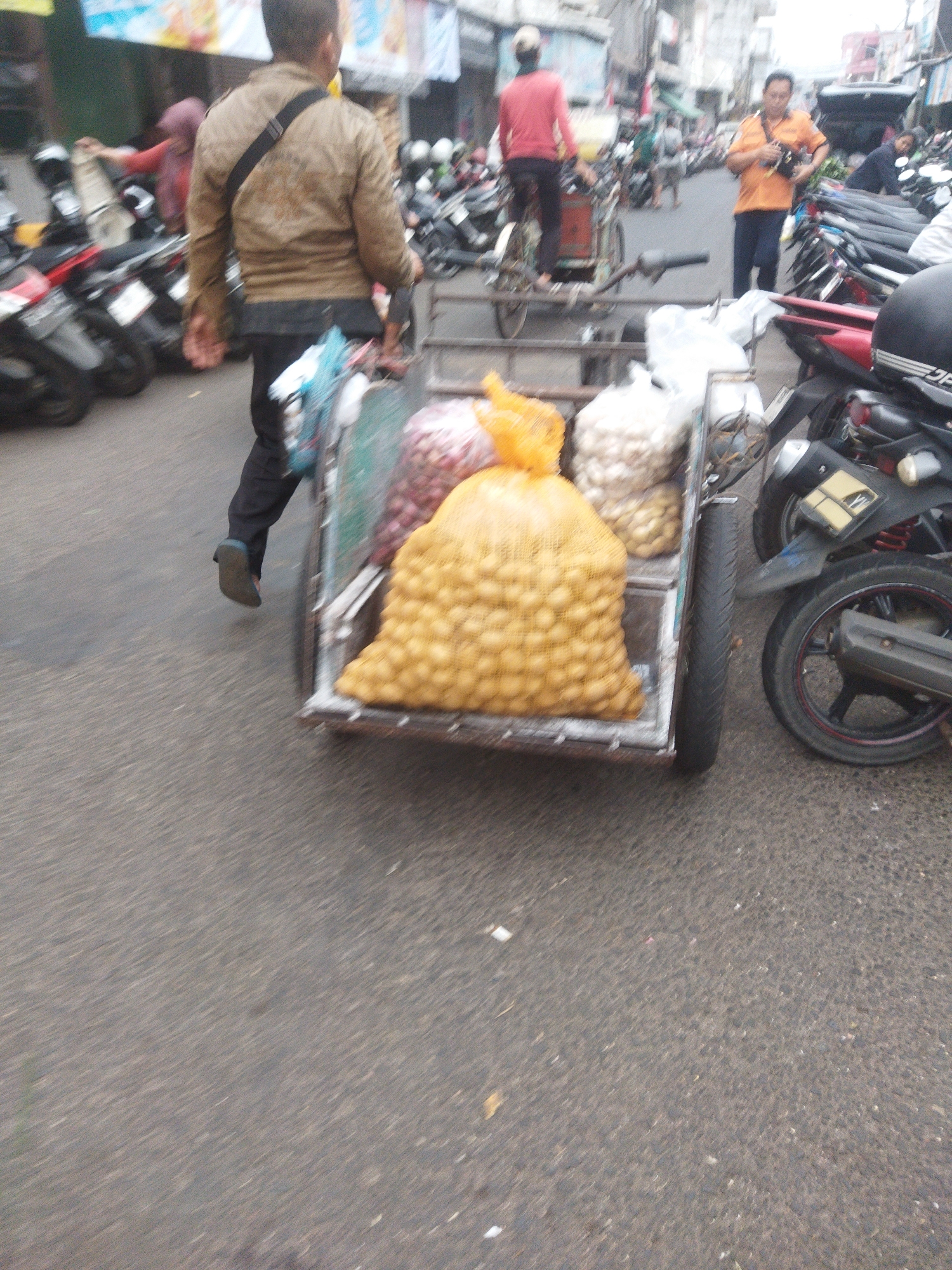 The image captures a bustling street scene, likely in Banjar, Indonesia, indicated by the presence of motorcycles, market stalls, and individuals engaged in commerce. In the foreground, a person wearing a light brown jacket and black pants pushes a cart laden with produce, primarily large sacks of potatoes and what appear to be onions or garlic. The cart is a two-wheeled, human-powered vehicle with a metal frame and a wooden bed.
Behind the cart, several motorcycles are parked or moving, suggesting a busy urban environment. To the left, a woman in a red headscarf and apron is visible, seemingly interacting with goods. Further down the street, other individuals are present, including a man in an orange shirt holding a camera, who appears to be a vendor or a photographer. The street itself is paved and shows signs of wear and tear, with some debris scattered on the surface. The background reveals market stalls with awnings and buildings lining the street, characteristic of a local marketplace. The overall impression is one of a vibrant, everyday commercial activity in a crowded Indonesian city.