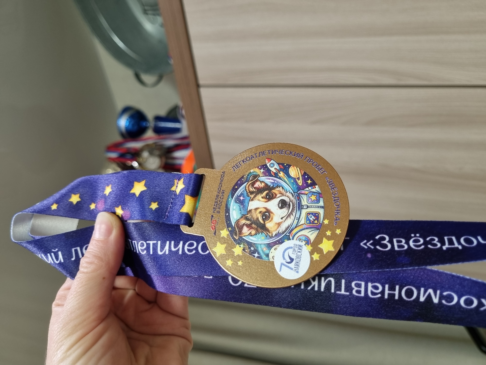 The image shows a medal held by a hand. The medal is golden and features a dog wearing a space suit in the center, surrounded by space-themed decorations. The ribbon attached to the medal is blue with a star pattern. The text on the medal indicates that it is related to a running race called "Star" and commemorates 75 years of the city of Chaikovsky, Russia.