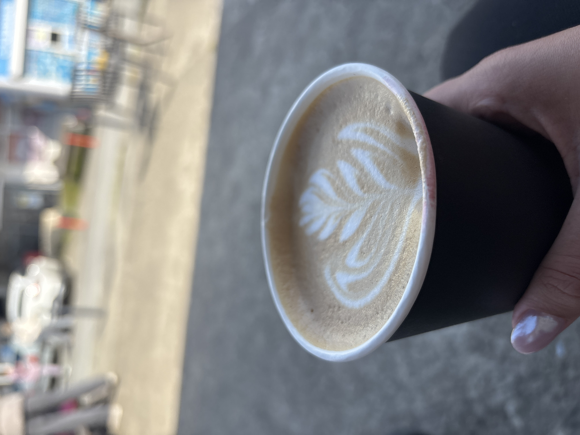 The image shows a cup of coffee held in a person's hand. The coffee has latte art, specifically a leafy pattern. The cup is black with a white rim. The background is blurred, but appears to be an outdoor urban setting. Based on the request for information about the city of Sochi, Russia, it is implied that the photograph might have been taken there. However, without additional information such as landmarks, signs, or architecture specific to Sochi, it is impossible to confirm the location with certainty.