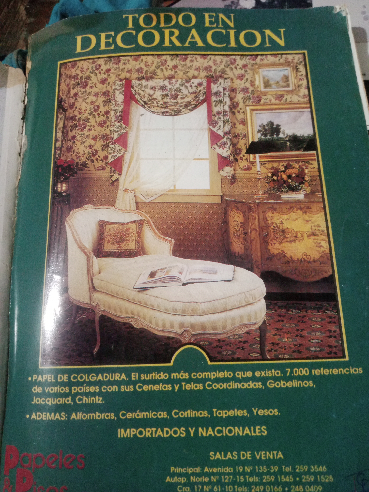 The image is a book cover titled "TODO EN DECORACION" (Everything in Decoration). The cover features a scene of a luxurious living room with floral wallpaper, a large window with ornate curtains, a vintage chaise lounge with a book on it, and a decorative cabinet with floral arrangements. The text indicates that the book offers a wide selection of wallpaper, borders, fabrics, rugs, and other decorative items. There is no information within the visible portion of the image that specifies the location within the city of Santa Ana, Venezuela. The image is a still life showcasing interior design elements.