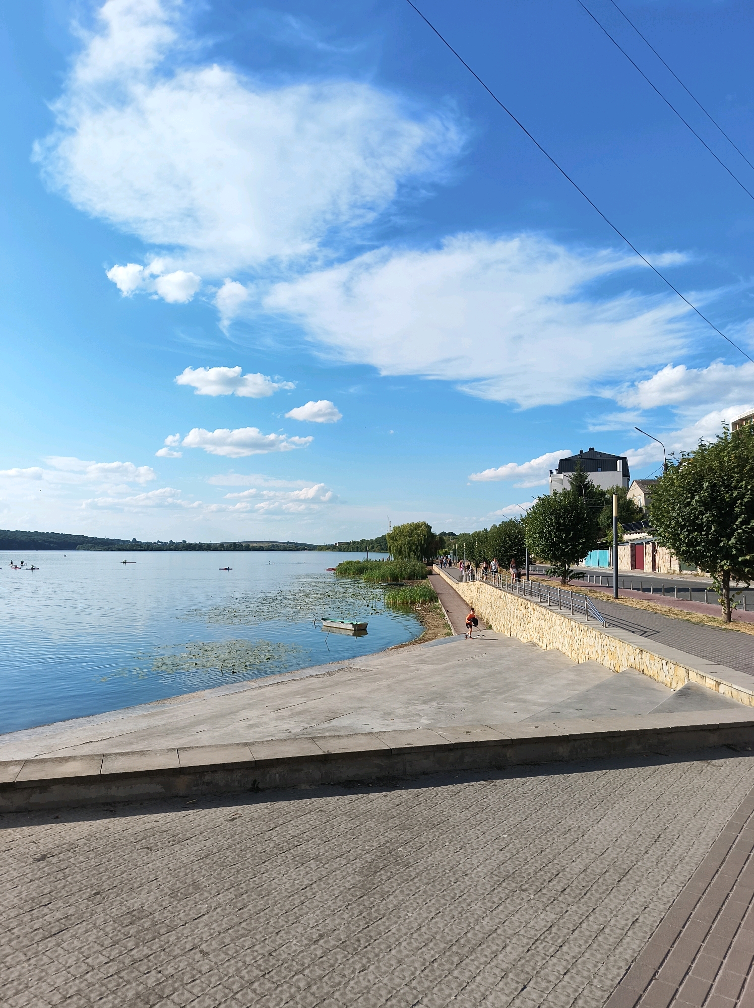 The image depicts a sunny day along the Ternopil Pond embankment, specifically near the "Natalka" embankment. The foreground is dominated by a textured concrete surface, a common feature of the renovated waterfront. Beyond this, a stepped concrete structure leads down to the water's edge. On the water, a small, light-colored boat with green trim is moored near a patch of water lilies. Several individuals are visible on the lake, some paddling kayaks or canoes, while others are in boats further out.
The embankment itself is a paved walkway lined with a low stone wall. A group of people are walking and cycling along the path, enjoying the day. Trees line the edge of the embankment, providing some shade. Buildings, including a multi-story structure with a dark roof, are visible behind the trees. The sky is a vibrant blue with scattered white clouds, indicative of pleasant weather. The overall scene conveys a sense of leisure and outdoor activity in a picturesque urban waterfront setting.