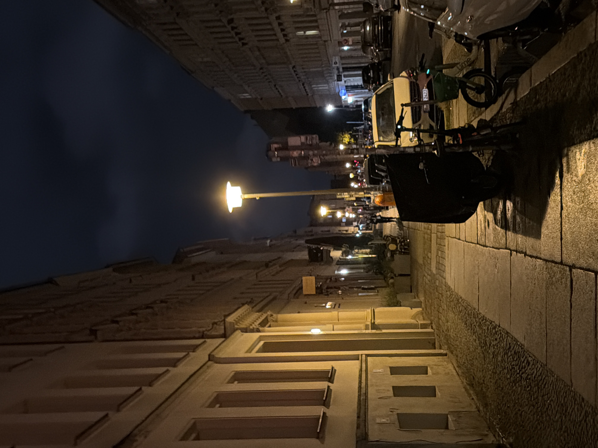 The image captures a street scene at night in Berlin, Germany, likely in a residential or mixed-use area with a historical architectural style. On the left, a light-colored building with multiple windows and a textured facade lines the street. The sidewalk is paved with flagstones and cobblestones, with a visible drainage grate. A streetlamp with a modern design illuminates the scene.
On the right side of the street, parked cars are visible, including a light-colored sedan and a darker SUV. Several bicycles and e-scooters are parked along the curb. Another building with multiple windows is visible on the right, also in a typical Berlin architectural style.
In the background, more streetlights and buildings recede into the distance. The sky is dark with some clouds, indicating nighttime. A few individuals are present on the sidewalk, walking. One person is seen standing near a table and chairs outside what appears to be a cafe or restaurant. The overall impression is that of a quiet urban street at night. The license plate of the light-colored car visible is B 2423.