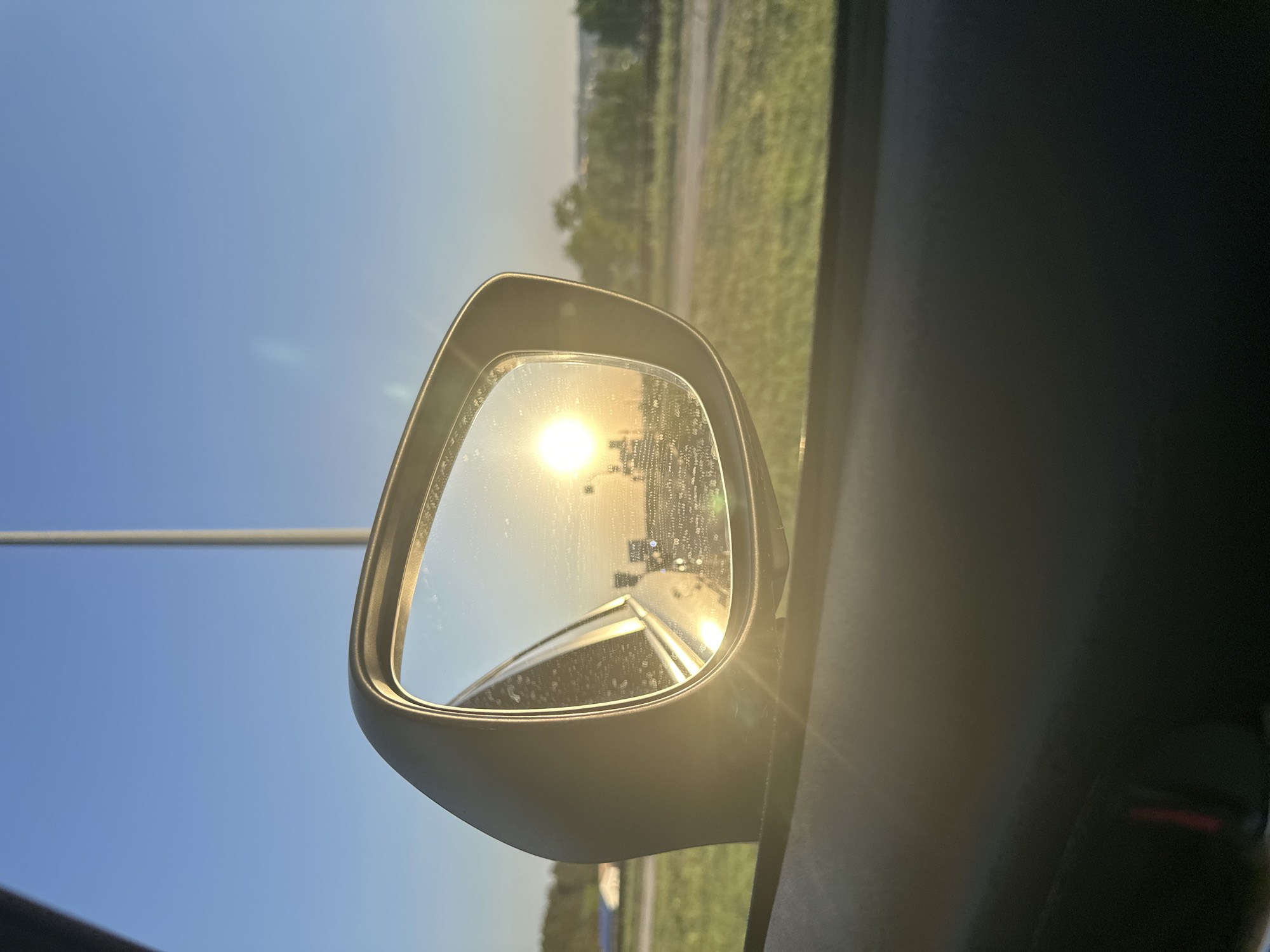 The image shows a side mirror of a car, reflecting the sun setting on a road in Chaikovsky, Russia. The mirror is mostly clean but has some water spots. The sun reflects brightly in the mirror, with other cars visible in the reflection. A concrete pole is visible outside the window on the left, while green grass and trees are on the horizon to the right, outside the mirror. The inside of the car, including the dark door panel, is visible on the right side of the picture.