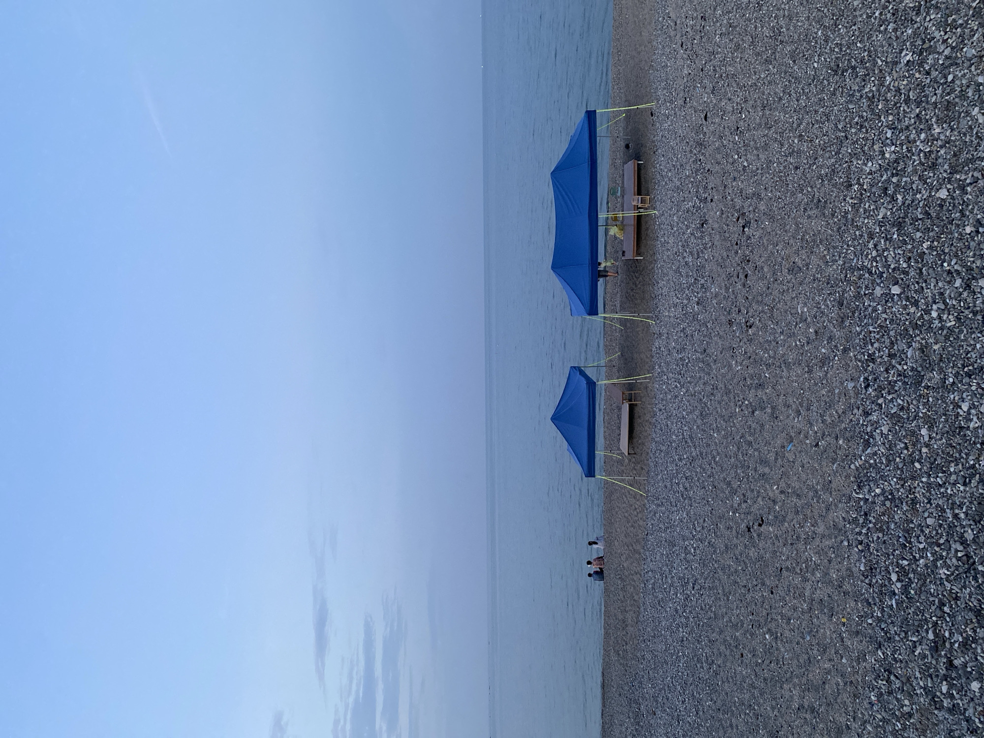The image depicts a serene beach scene at dusk, likely in Gyeongju-si, South Korea, given the context provided. The sky is a pale blue, transitioning to a darker hue towards the horizon, with wispy clouds scattered across the upper half of the frame. The sea is calm and stretches out to meet the sky, reflecting the twilight.
In the foreground, the beach is covered in pebbles and sand. Two blue canopies are set up on the beach, providing shade. Under the canopies are simple wooden benches, suggesting a place for relaxation or a temporary setup for an event. One canopy has a table and chairs beneath it.
To the left of the canopies, three people are sitting on the edge of the beach, facing the sea, appearing to be engaged in conversation or simply enjoying the view. Their postures suggest a relaxed and leisurely atmosphere. One person under the right canopy appears to be standing. The overall impression is one of tranquility and peacefulness as day transitions into night.