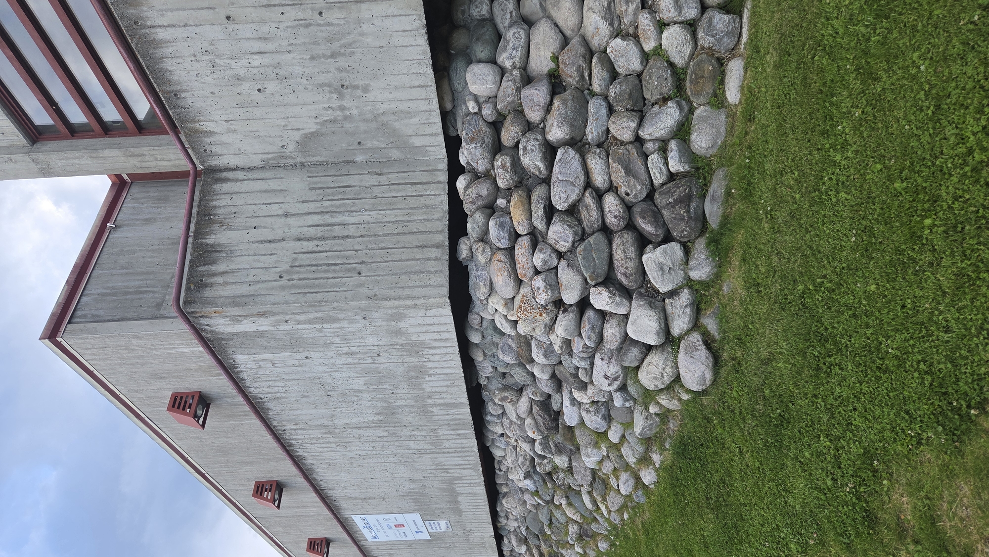 The image shows the exterior of a modern concrete building with a stone foundation. The building has a red trim along the roofline and around the windows. There are three red outdoor light fixtures visible on the side of the building. A sign is mounted on the concrete wall. The stone foundation is constructed of various sized, rounded river stones. In front of the stone foundation, there is a well-maintained green lawn. The sky is overcast with clouds. The location is identified as Tynset, Norway.