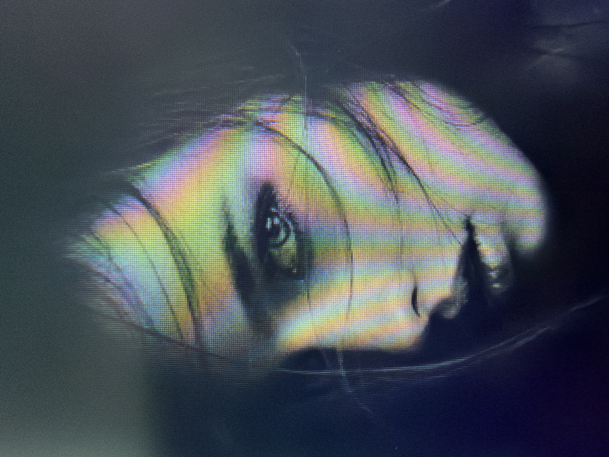 The image is a close-up, low-resolution, pixelated photograph of a woman's face, seen from the shoulders up, in a dark, atmospheric setting. Her hair is windswept across her face, obscuring parts of her features. The lighting is dramatic, with strong highlights and deep shadows, creating a sense of mystery and intensity. The woman's eyes are visible and appear to be looking slightly off-camera. Her lips are slightly parted. The overall tone of the image is somber and artistic. There is no discernible location in Plovdiv, Bulgaria, or any other city. The image depicts only the woman's face and does not show any environmental context.
