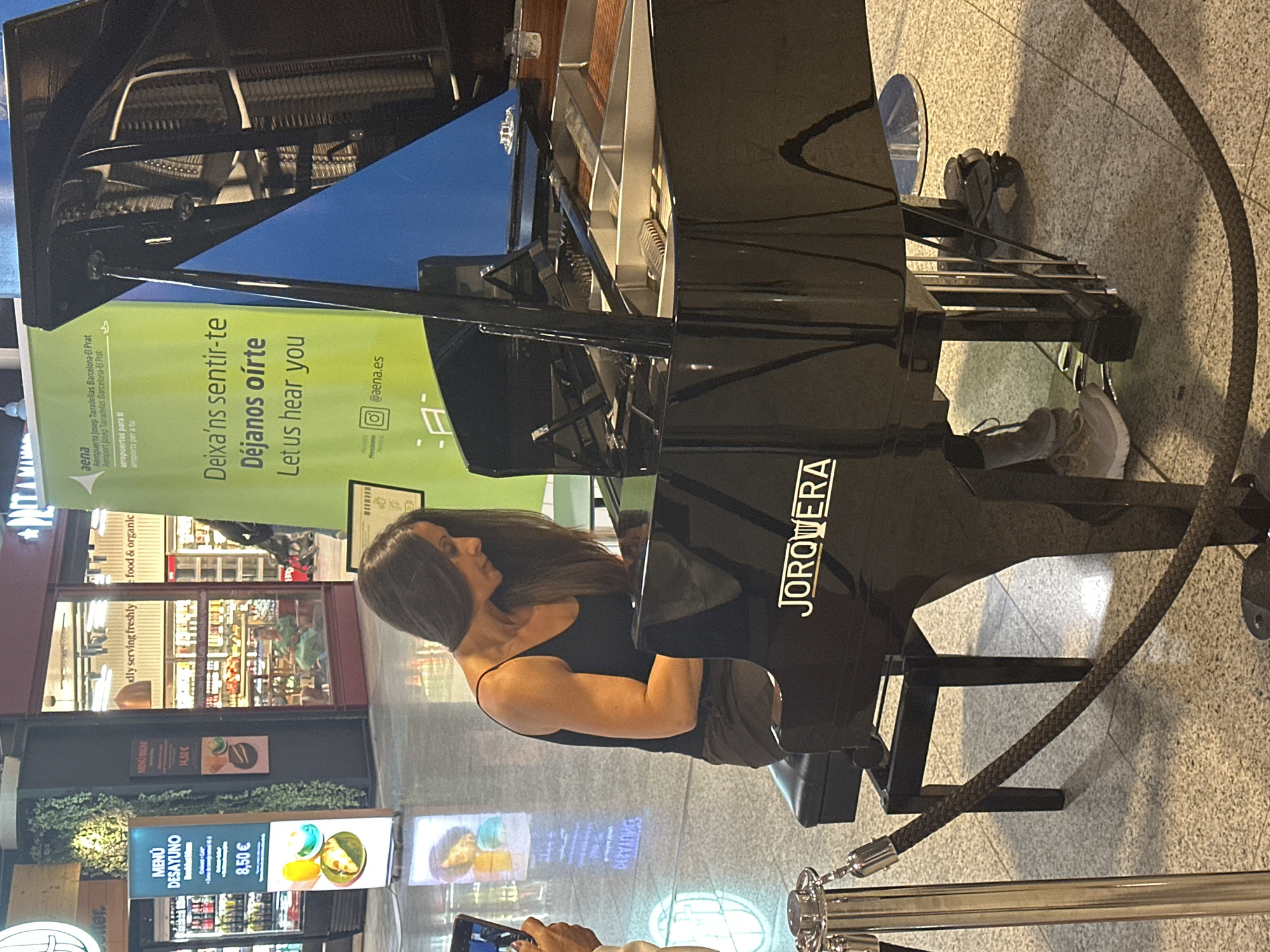 The image captures a woman playing a grand piano in a public space, likely an airport terminal at El Prat de Llobregat, Spain, suggested by the presence of AENA branding on a nearby banner. The piano is black and appears to be a performance instrument, with "JORQWERTY" visible on its side. The woman, wearing a black tank top and gray pants, is seated at the piano, her hands on the keys, and her feet on the pedals. Her gaze is directed downwards at the keyboard. The background includes a green banner with text in Catalan, Spanish, and English, promoting Aena and its airports. Other elements visible are stanchions with rope barriers, reflective flooring, and storefronts in the distance, typical of an airport concourse. The lighting suggests an indoor, well-lit environment.