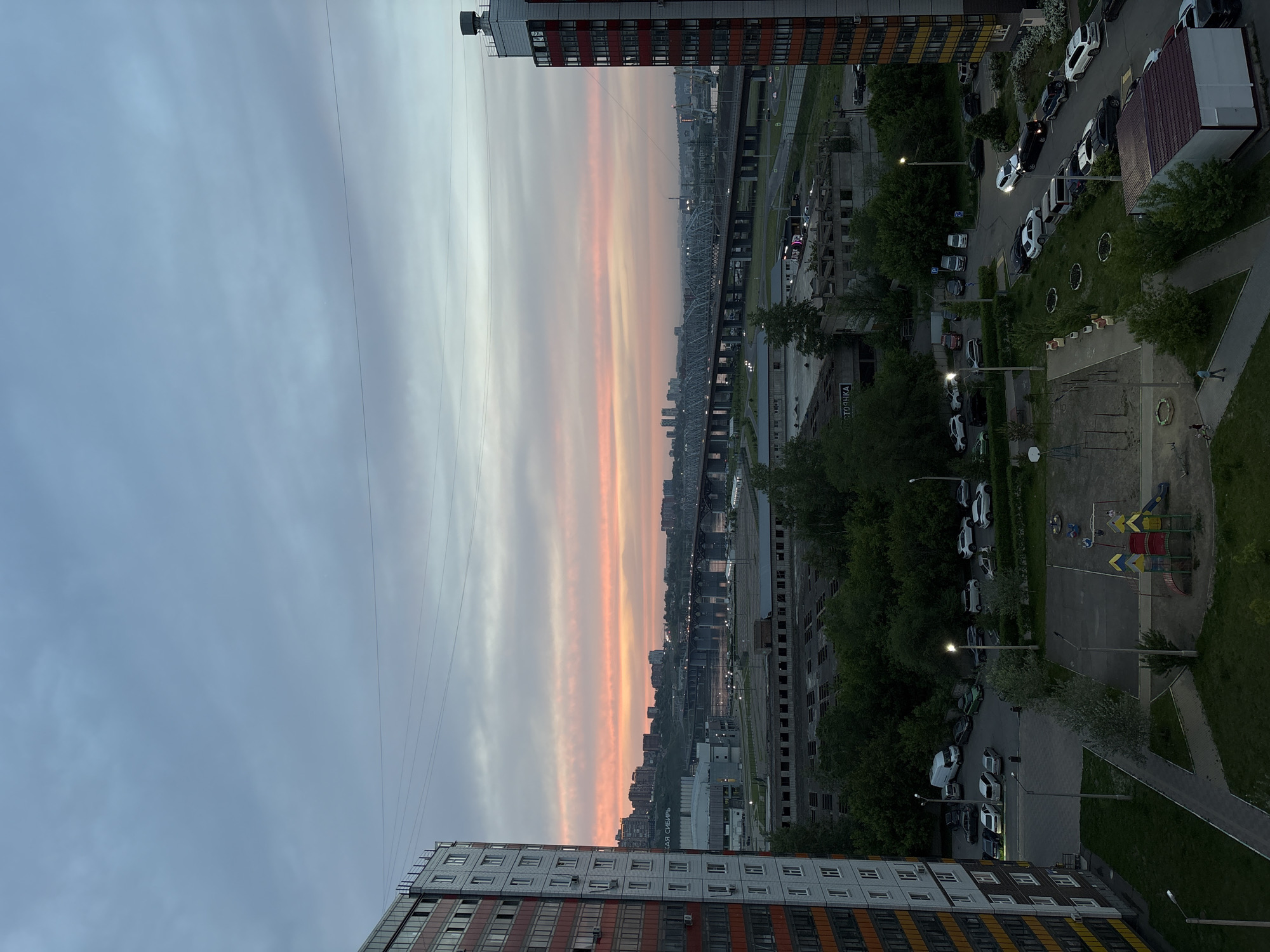 This is a vertical, eye-level, daytime, medium shot of a city skyline at sunset, with a focus on the sky and an apartment building. The sky is filled with dramatic clouds in shades of gray, white, and orange, creating a striking sunset. In the foreground, a modern apartment building with a red and orange facade is visible, with many windows reflecting the sky. In the middle ground, a river and a bridge are seen, with a city skyline in the distance. The overall mood is peaceful and serene, with a hint of urban beauty. The lighting suggests it is either early morning or late evening due to the soft, diffused light and the sunset colors.