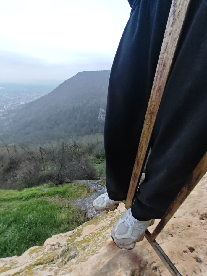 An elevated perspective captures the lower legs and feet of an individual, dressed in black pants and white and grey athletic shoes, positioned on a light-colored, rocky ledge. A rusty, brown metal frame or ladder is positioned vertically in the immediate foreground, partially obscuring the individual's right leg. Patches of green grass and moss are present on the rocky surface to the left. The scene looks downward over a steep, forested slope featuring numerous bare deciduous trees and a prominent grey rock face on the right midground. In the distant background, the urban landscape of the city of Shumen, Bulgaria, is visible, partially obscured by atmospheric haze. The sky is overcast and uniformly pale grey.