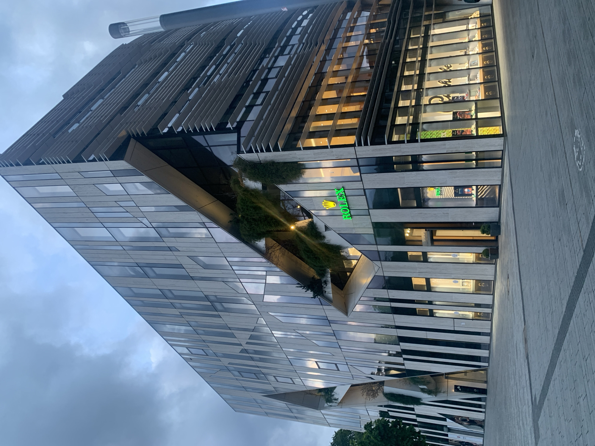 This image captures the Kö-Bogen II building in Düsseldorf, Germany, under an overcast or twilight sky. The modern, multi-story structure dominates the frame, showcasing a distinctive architectural design. The building's facade is characterized by alternating vertical panels of reflective glass and light-colored material, creating a dynamic visual pattern. On the left side, the building features large, angular cutouts that house integrated vertical gardens with lush green vegetation, illuminated from within. The right side of the building presents a curved or rounded facade, accented by multiple horizontal louvers or fins across its upper stories.
At ground level, the building houses retail establishments with expansive window displays. A prominent, illuminated green "ROLEX" sign with its crown logo is visible on the left side of the ground floor. Adjacent to it, further along the facade, are numerous window displays showcasing luxury watches and jewelry. The name "Bucherer" is partially visible on one of these store windows, identifying a luxury retailer. The interior lights of these stores are active, casting a warm yellow glow onto the merchandise and pavement, contrasting with the cooler ambient light.
The foreground consists of a wide, clean paved plaza or walkway composed of light and dark grey rectangular paving stones. A dark linear strip is embedded within the paving. To the far right of the building, a tall, slender, dark cylindrical structure with a transparent, illuminated top section stands vertically. The sky is largely overcast, displaying varying shades of grey and muted blue. Trees are partially visible on the far left edge of the frame. No individuals are prominently visible in the immediate foreground.