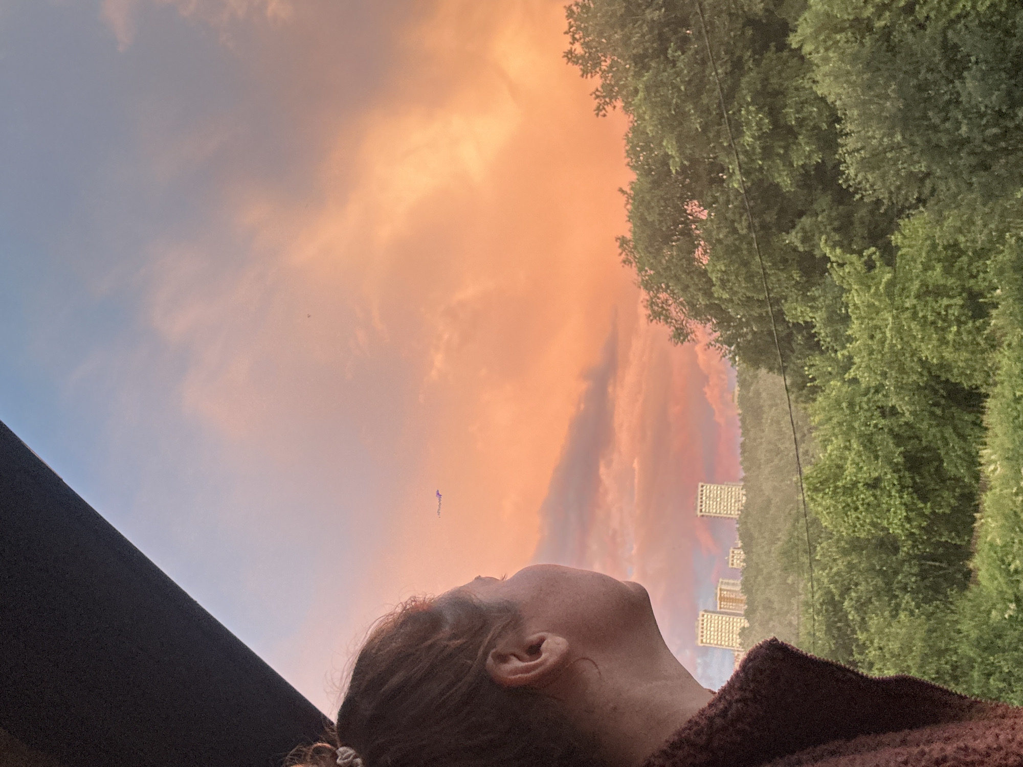 A woman is pictured from the side, her face turned towards a vibrant sunset. Her hair is pulled back in a bun, and she is wearing a dark, textured garment. In the background, a cityscape emerges from a verdant, tree-covered hillside. The sky is a dramatic canvas of orange, pink, and blue, with soft clouds stretching across it. A small airplane is visible in the distance, a dark silhouette against the colorful sky. The overall scene evokes a sense of tranquility and contemplation, with the natural beauty of the sunset juxtaposed against the urban landscape. This vista is likely from a high vantage point, possibly a balcony or rooftop, offering an expansive view of Nizhny Novgorod, Russia.