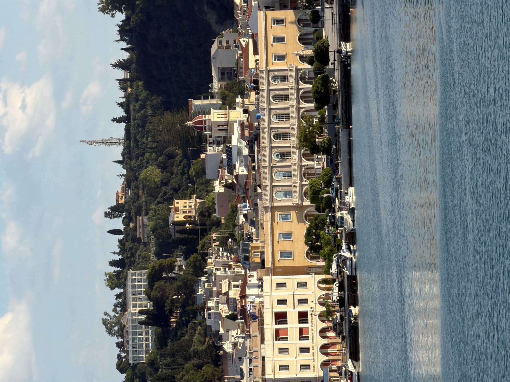 The image captures a waterfront scene in Zakynthos Municipal Unit, Greece, showcasing a harbor filled with boats and a backdrop of buildings and a lush, verdant hillside. The water in the foreground is a tranquil blue, reflecting the light. Along the waterfront, several boats of varying sizes are docked, some appearing to be small pleasure crafts. Behind the boats, a row of buildings with arched ground floors and multiple stories extends along the harbor. These buildings exhibit a mix of architectural styles, with some featuring balconies and a variety of window designs. One prominent building is a light yellow, with a colonnaded facade on its upper floors and arched doorways at street level. To the right of this building, a church with a red-domed bell tower rises above the lower structures, its spire reaching towards the sky.
The hillside behind the buildings is densely covered with trees and vegetation, creating a rich green backdrop. Various buildings, including what appears to be a large hotel or complex with numerous windows, are scattered across the hillside, some situated at higher elevations. A prominent communication tower stands on the crest of the hill. The sky above is a bright blue with scattered white clouds, suggesting a clear day with ample sunlight, which casts shadows and highlights the architectural details. No people are clearly discernible in action or interaction, though a few figures might be present near the waterfront buildings. The scene conveys a sense of a peaceful coastal town.