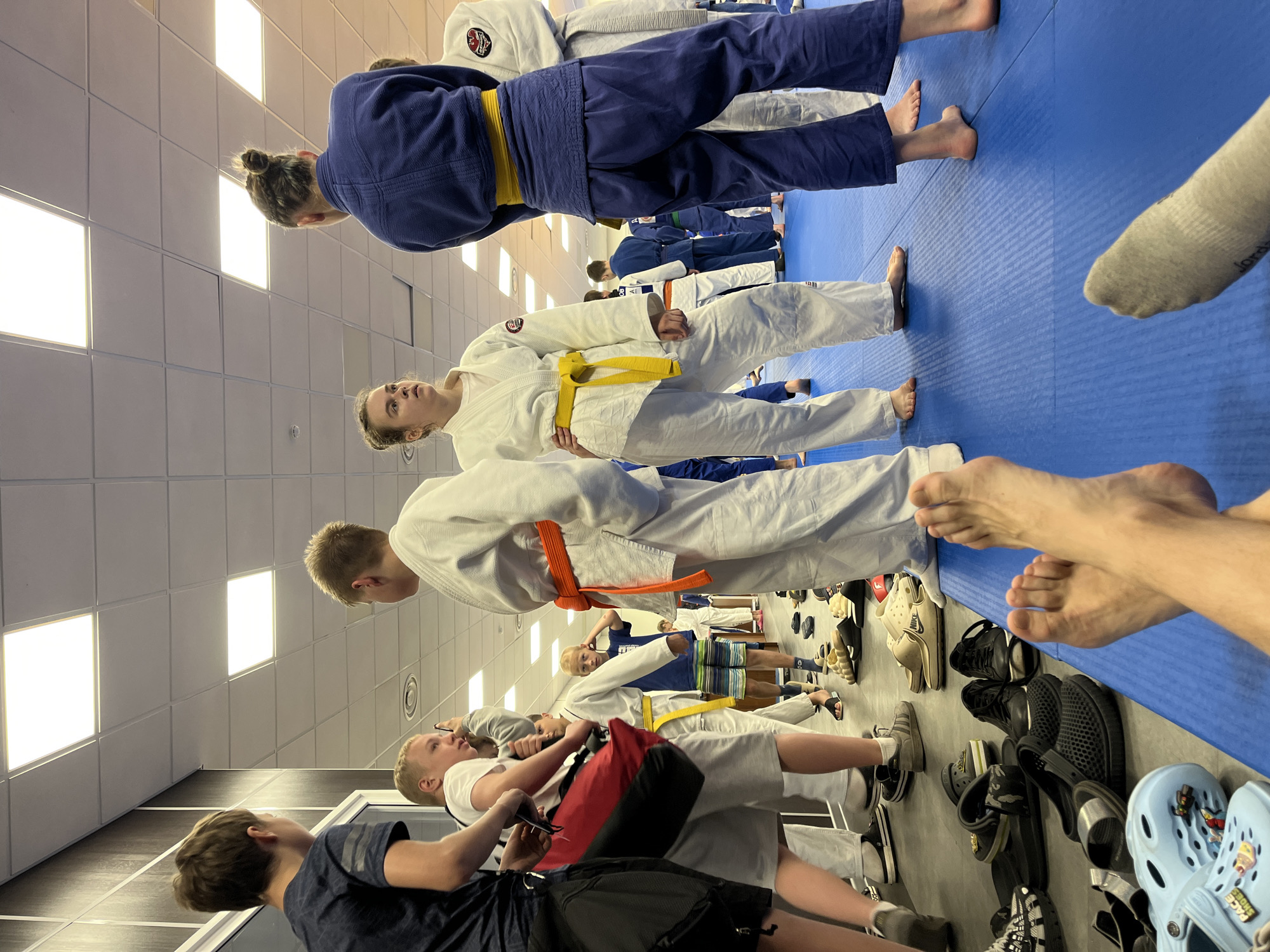The image depicts an indoor martial arts training facility, specifically a dojo, located in Krasnoyarsk, Russia. The scene captures a group of children and young adults, most of whom are dressed in judogi (martial arts uniforms) of either white or blue, indicating participants in a judo or similar grappling sport class. The floor is covered with blue, textured mats designed for martial arts training.
In the foreground, from a low perspective, are the bare, crossed feet of one individual, while another foot, partially clad in a grey sock, is visible in the lower right. This suggests the viewpoint of someone resting on the mat.
In the mid-ground, multiple individuals are standing. On the far left, a male youth in a dark blue t-shirt and dark shorts holds a red bag. Adjacent to him, another male youth in a white t-shirt and grey shorts appears to be adjusting something near his head. Centrally, a male youth wears a white judogi with a bright orange belt, facing right. Next to him, a female youth in a white judogi with a yellow belt looks upwards and to the left. Behind her, a female youth in a blue judogi with a yellow belt is seen from behind, her hair in a bun. To her right, another individual is seen from behind in a white judogi, featuring a dark circular emblem on the left chest.
In the background, other participants, some in judogi and some in casual attire like a blue t-shirt and striped shorts, are visible standing or moving within the space. A significant pile of assorted footwear, including sneakers, sandals, Crocs, and slides, is gathered off the mat along a wall, indicating the designated area for shoes.
The facility features light-colored walls, with some darker paneling visible on the left. The ceiling is a suspended grid system with multiple large, rectangular LED light panels providing bright illumination. Circular ventilation grilles are also visible on the ceiling. The overall atmosphere suggests an active training environment or a brief pause during a session.