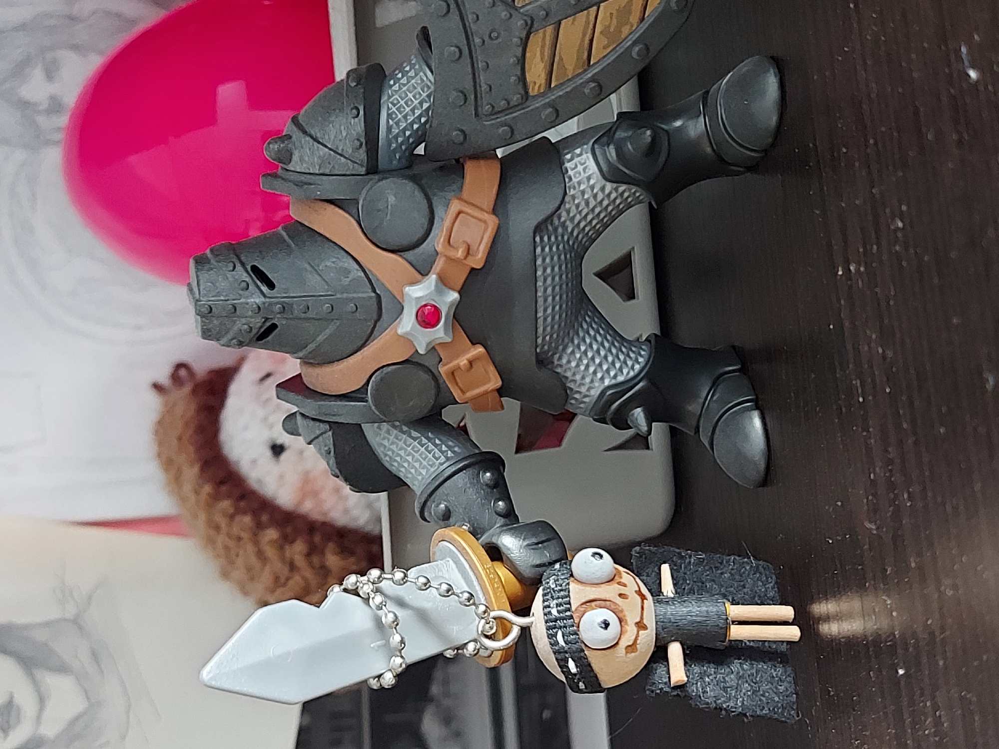 The image shows a toy figurine of a medieval knight pig with a sword and shield. It is lying on a dark surface, possibly a wooden table. A small keychain figure of a crude human-like character with large eyes lies near the pig figurine. In the background, there is a pink object, a crocheted hedgehog, and other unidentifiable items. The location of the image is unknown and there is no indication it is taken in the city of Izhevsk, Russia.