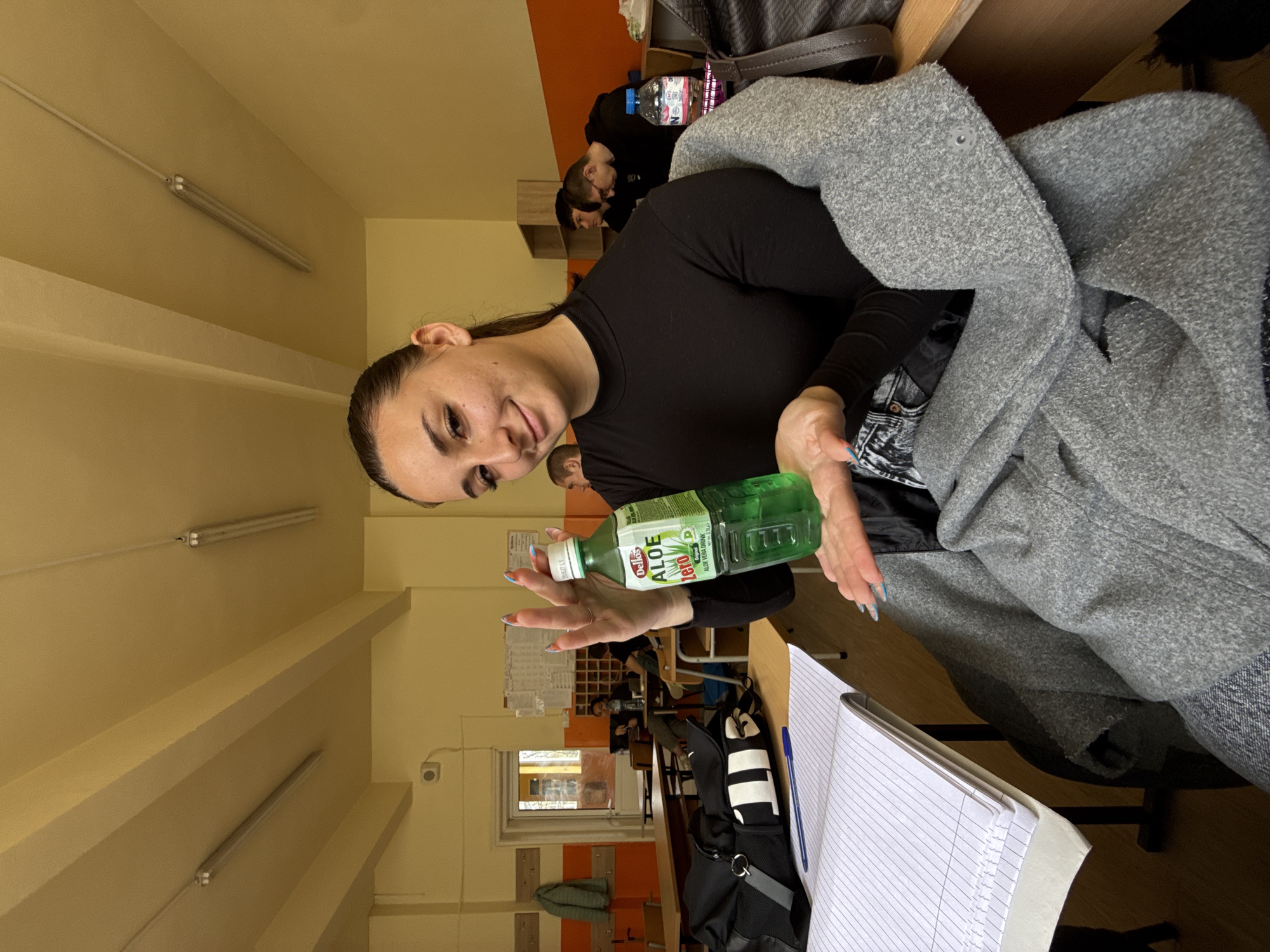 The image depicts an indoor scene, likely a classroom or lecture hall. A young woman is prominently featured, holding a bottle of "Dellos Aloe Zero" aloe vera drink. She is wearing a black long-sleeved shirt and has her hair pulled back. A grey sweater or jacket is draped over a chair or nearby surface.
Other individuals are visible in the background, seated at desks, suggesting an educational setting. A notebook and pen are on the desk in the foreground, adding to this impression. The room has yellow walls and fluorescent lighting. Through a doorway, a hallway can be seen.
The location is potentially Sofia, Bulgaria, but that would require additional information or context to verify. The style of architecture and interior design is consistent with buildings found in that region. The presence of a product like "Dellos Aloe Zero" provides further geographical context, as this would indicate the image was likely taken in a location where this product is available.