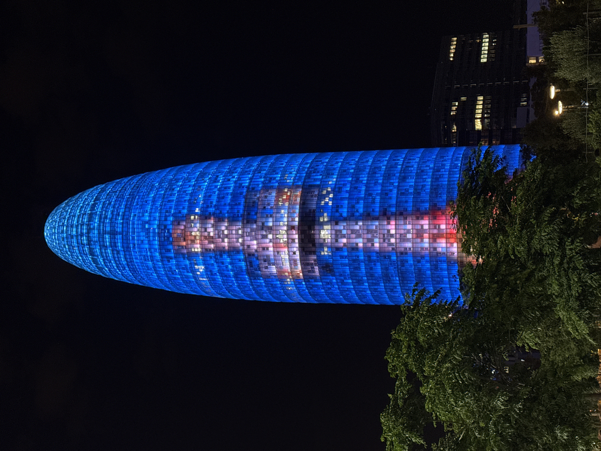 The image shows the Agbar Tower in Barcelona, Spain, at night. The skyscraper, illuminated with blue and red lights, features a mosaic pattern of lights on its facade that resembles a human figure. Trees and other buildings are visible in the foreground and background, respectively, with streetlights illuminating the lower portion of the scene. The sky is dark, indicating nighttime.