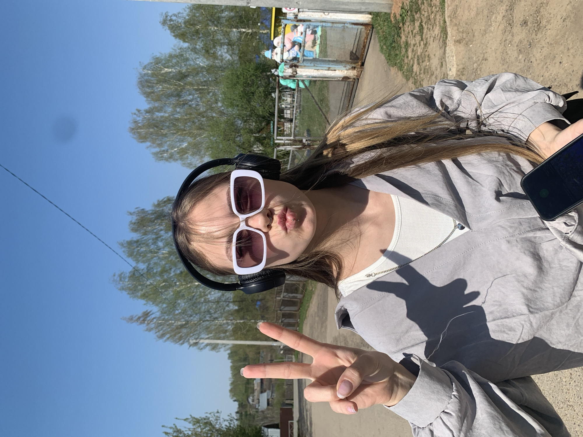 The image depicts a young woman outdoors, likely in the city of Фоки, Russia. She is wearing sunglasses and headphones and making a peace sign with her right hand. She has light brown hair and is wearing a grey jacket over a white top. She is holding a smartphone in her left hand. The background includes trees, a blue sky, and a fenced structure that appears to be a playground. The ground is a mix of concrete and dirt.