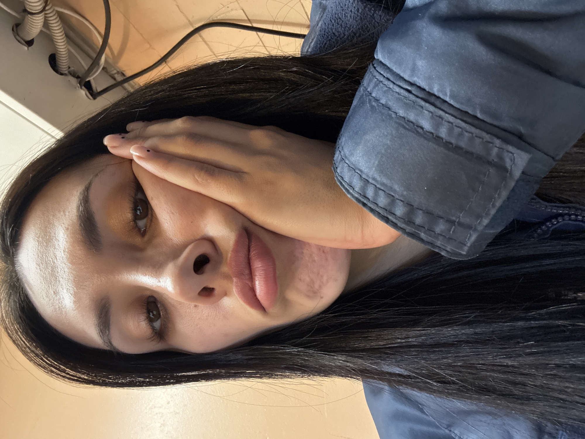 A close-up portrait, taken in Votkinsk, Russia, features a person, likely a young woman, with long, straight dark hair. The image is presented on its side, showing her head significantly tilted. Her dark brown eyes gaze directly at the viewer. Her left hand is pressed against the left side of her face, partially obscuring her cheek and mouth. Her fingernails have a light polish adorned with small black dots. Her lips are full and slightly pursed in a pout, with some skin texture or marks visible on her lower left cheek.
She is wearing a dark blue jacket, visible on the right side of the frame, showing stitched details and a textured fabric. Her long hair cascades around her face and over her shoulder, blending with the jacket. The background is simple, consisting of a plain, light brown wall. In the upper left corner, utility elements such as corrugated pipes, electrical wires, and a white fixture are visible against a lighter-colored surface, suggesting an indoor, possibly utilitarian setting. The lighting appears to be artificial. The subject's expression seems pensive or possibly playful. No text is visible in the image.