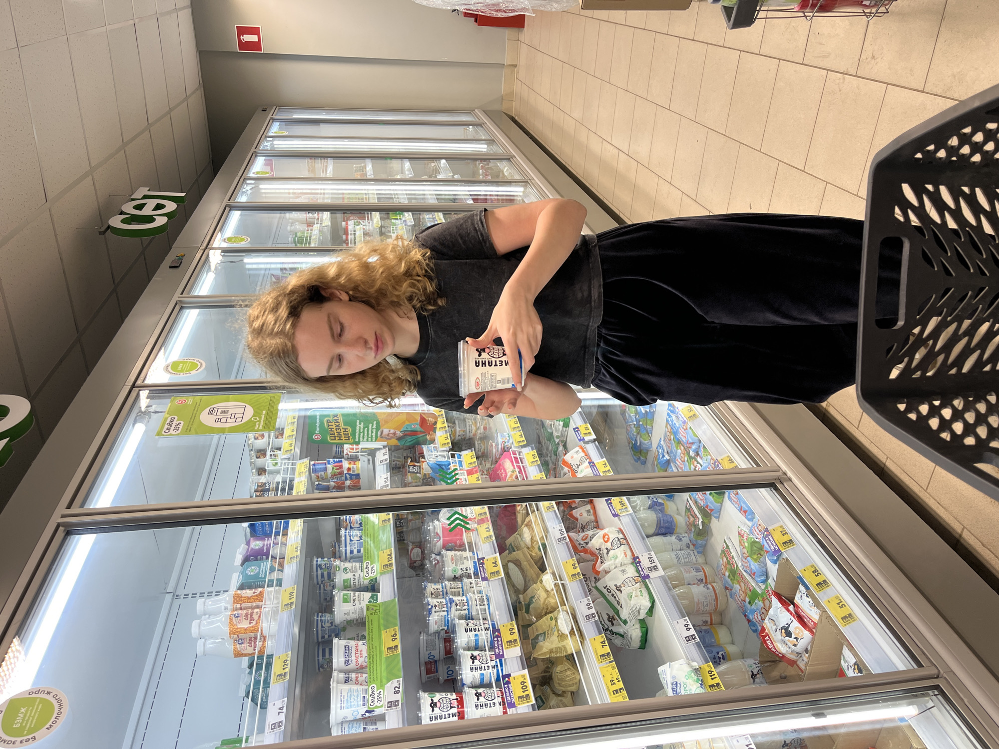A young woman with curly blonde hair is standing in an aisle of a supermarket, holding a container of yogurt. She is wearing a dark t-shirt and dark pants, and has a shopping cart in front of her. The supermarket aisle is lined with refrigerated display cases filled with dairy products. Signs with prices and discounts are visible on the shelves. The store appears to be a "Set' supermarket, a chain found in Russia, including in the city of Vasilyevo. The floor is tiled, and the lighting is bright. The woman is looking down at the yogurt container.