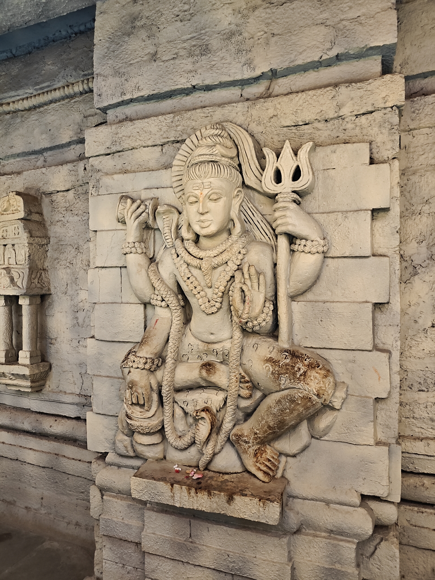 The image features a high-relief stone sculpture of a multi-armed Hindu deity, likely Shiva, carved into a light-colored, rough-hewn stone wall. The sculpture is rendered in shades of white and light grey, with some darker patinas and dirt, especially on the lower portions.
The deity is depicted in a seated or crouched posture, with one leg bent and the foot resting on the base, and the other knee raised. Its face is serene, bearing a prominent third eye mark (tilak) on the forehead. Adornments include multiple beaded necklaces, bracelets, and a prominent serpent coiling around the torso and left arm. In one of its right hands, the deity holds a damaru (small drum), while another right hand is raised in the Abhaya Mudra, a gesture of blessing or fearlessness. A trishula (trident) is firmly grasped in a left hand, extending vertically.
The surrounding wall is constructed from irregular stone blocks, displaying a textured surface and visible cracks, indicating age or weathering. To the left of the main sculpture, a smaller, intricately carved stone pillar or miniature shrine stands, featuring multiple tiers and decorative motifs.
At the base of the main sculpture, small offerings, including petals and other ritual items, are visible, suggesting an act of worship. The lighting suggests daylight. The scene conveys a strong sense of spiritual reverence within a temple or religious complex, aligning with the provided location context of Manikaran, India. No legible text is discernible on the sculpture or the surrounding architecture.