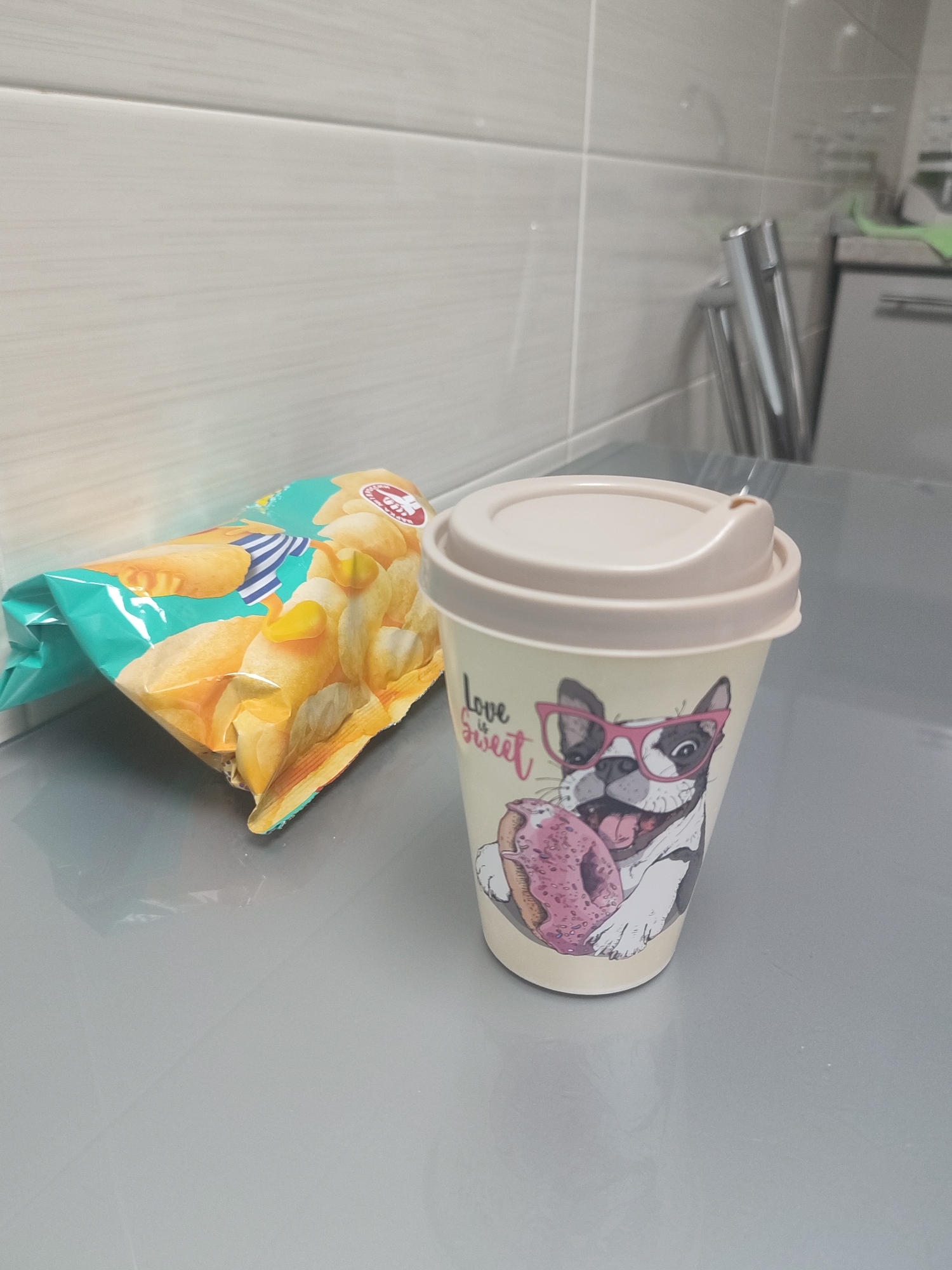 A plastic travel mug with a beige lid featuring a cartoon illustration of a French bulldog wearing pink glasses and eating a donut is placed on a grey countertop. Beside the mug, a partially opened bag of potato chips with a teal and yellow design rests on the counter. The background consists of light grey tiled walls. This scene appears to be located in a kitchen or break room, not specifically identifiable as Moscow, Russia, or any particular city.