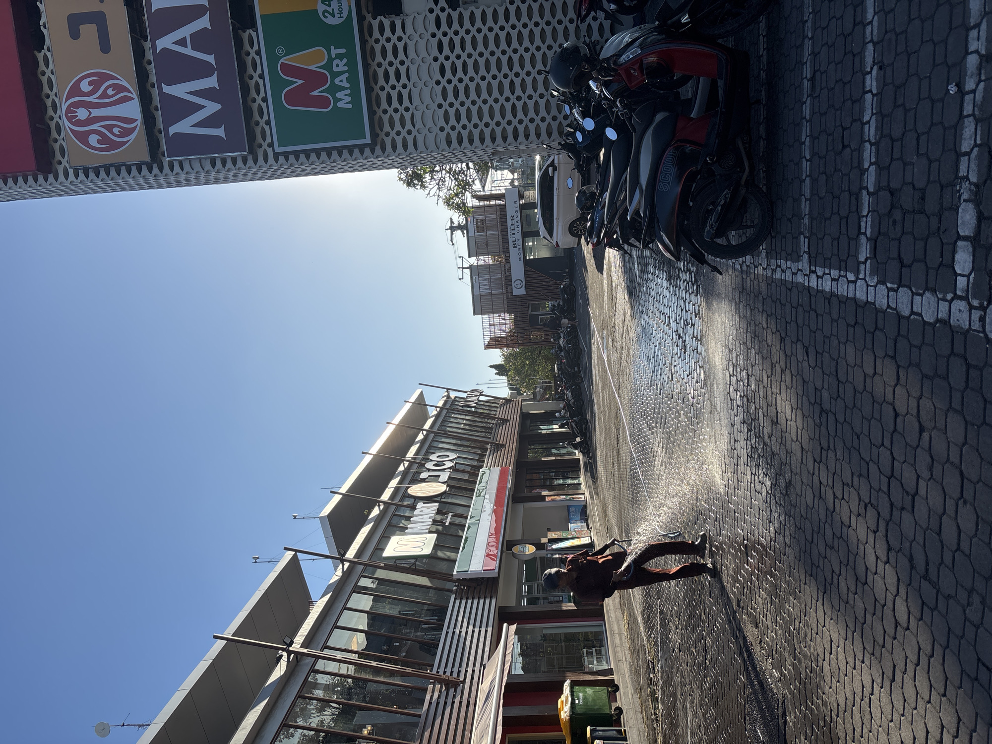 Here is a detailed description of the image:
**Location:** The image depicts a street scene in Kuta Selatan, Indonesia. The sky is clear blue.
**Objects and Subjects:** The image shows a street in front of commercial buildings, including a "M Mart" convenience store and a "Butler Money Changer". Motorbikes are parked along the side of the road. A man is actively cleaning the road with a water sprayer. A trash bin is also visible.
**People and Actions:** A man is cleaning the pavement with water.
**Environment:** The road is paved with cobblestone-like bricks. The scene is bathed in sunlight.