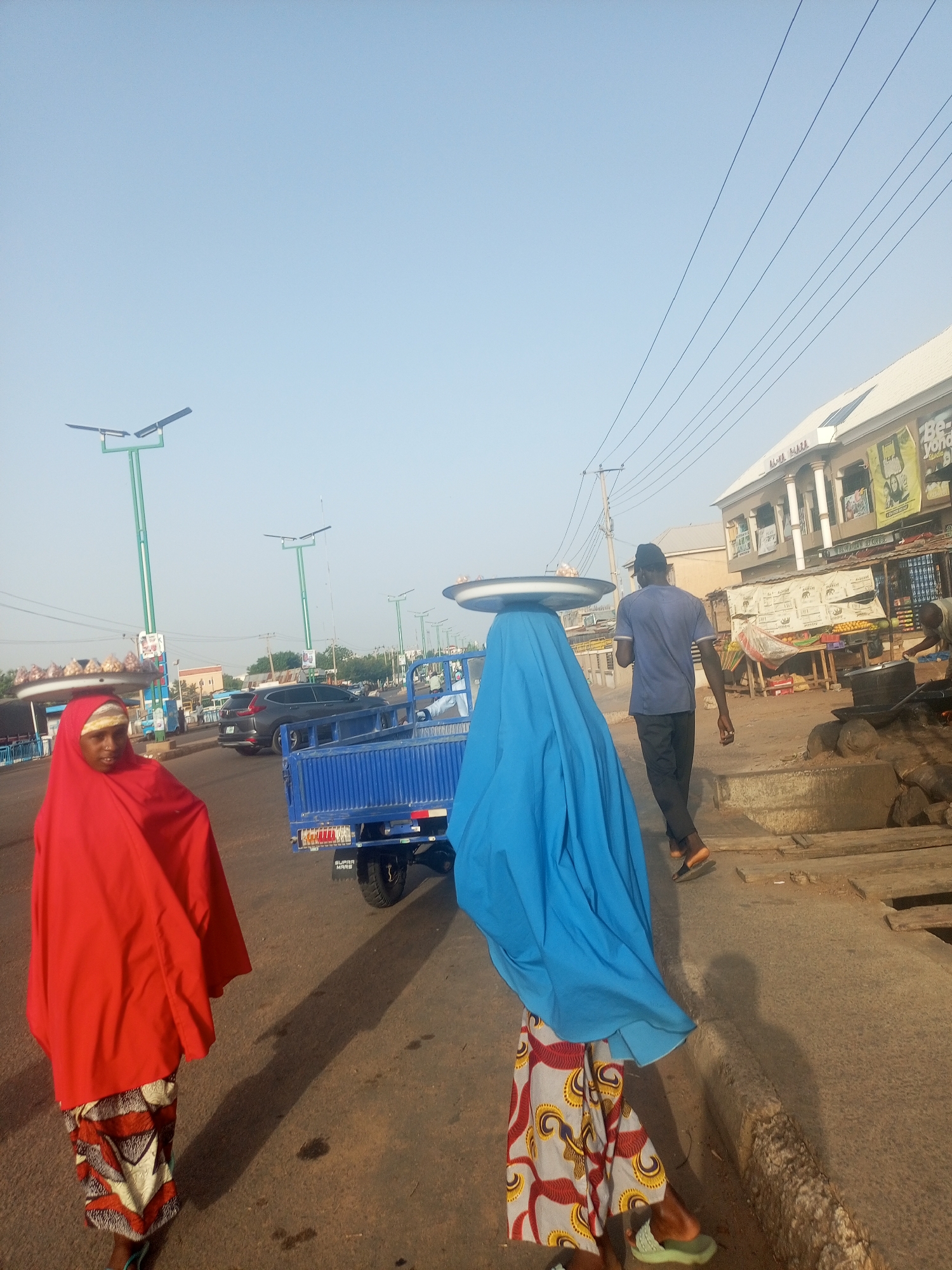 The image captures a street scene in Gombe, Nigeria. Two women are walking along the road's edge, each covered in brightly colored fabric. The woman on the left is draped in red and the woman on the right is draped in blue. Both are wearing patterned skirts and balancing trays on their heads. A man wearing a blue shirt and black pants is walking in the same direction behind them, also carrying a tray on his head. A small blue truck is parked on the road behind the women. A two-story building is visible on the right, and a person is seen cooking food outside. Solar-powered street lights line the road. The sky is clear and bright.