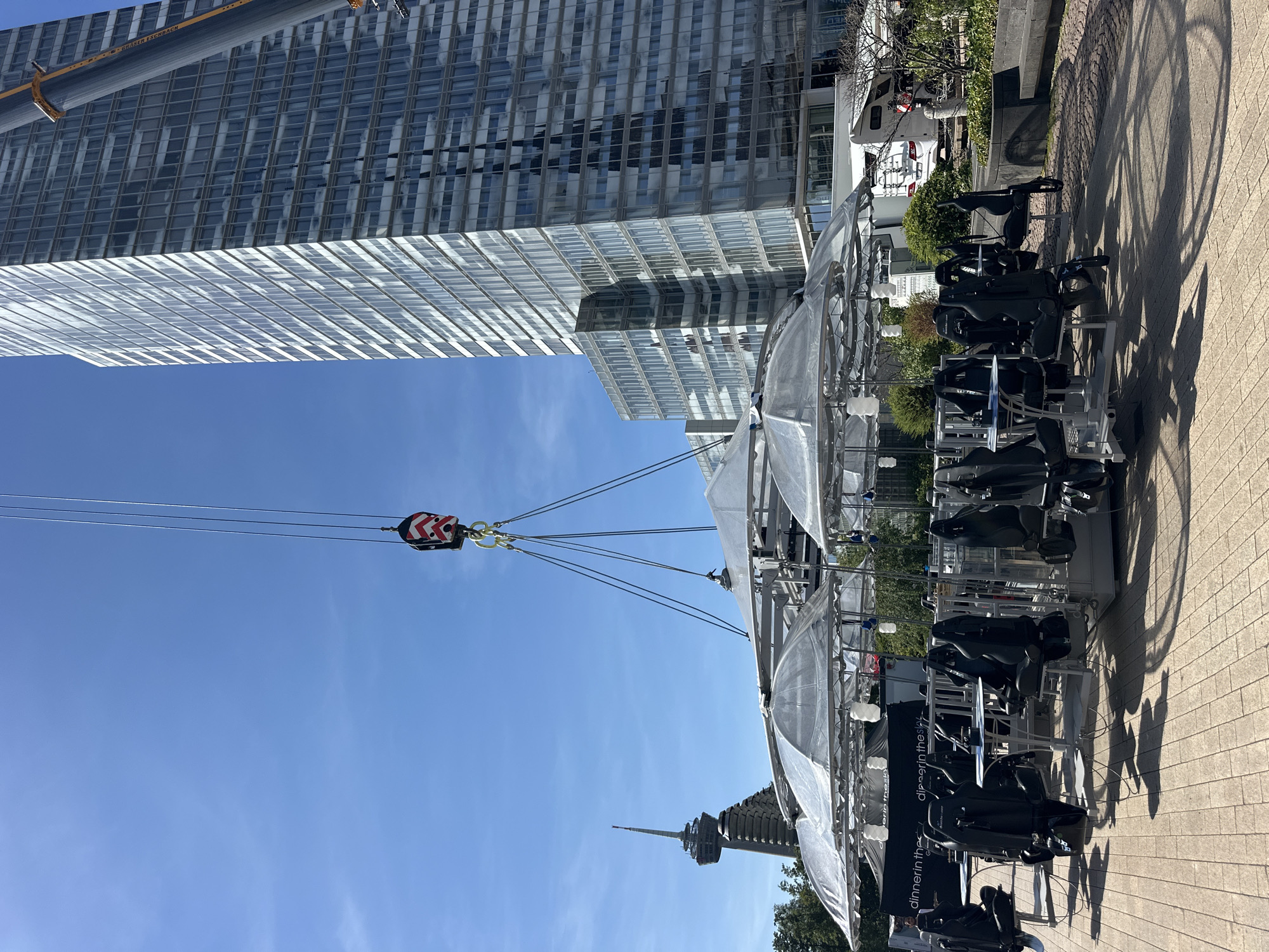 A crane is lifting a circular dining platform with attached seats and tables suspended in the air. The platform is covered with transparent umbrellas. In the background, a tall, modern glass skyscraper, the Colonia Tower (Colonius), and other buildings are visible under a clear blue sky. The scene appears to be in an urban outdoor setting, possibly a public square or park. The dining platform setup is the "Dinner in the Sky" attraction.
