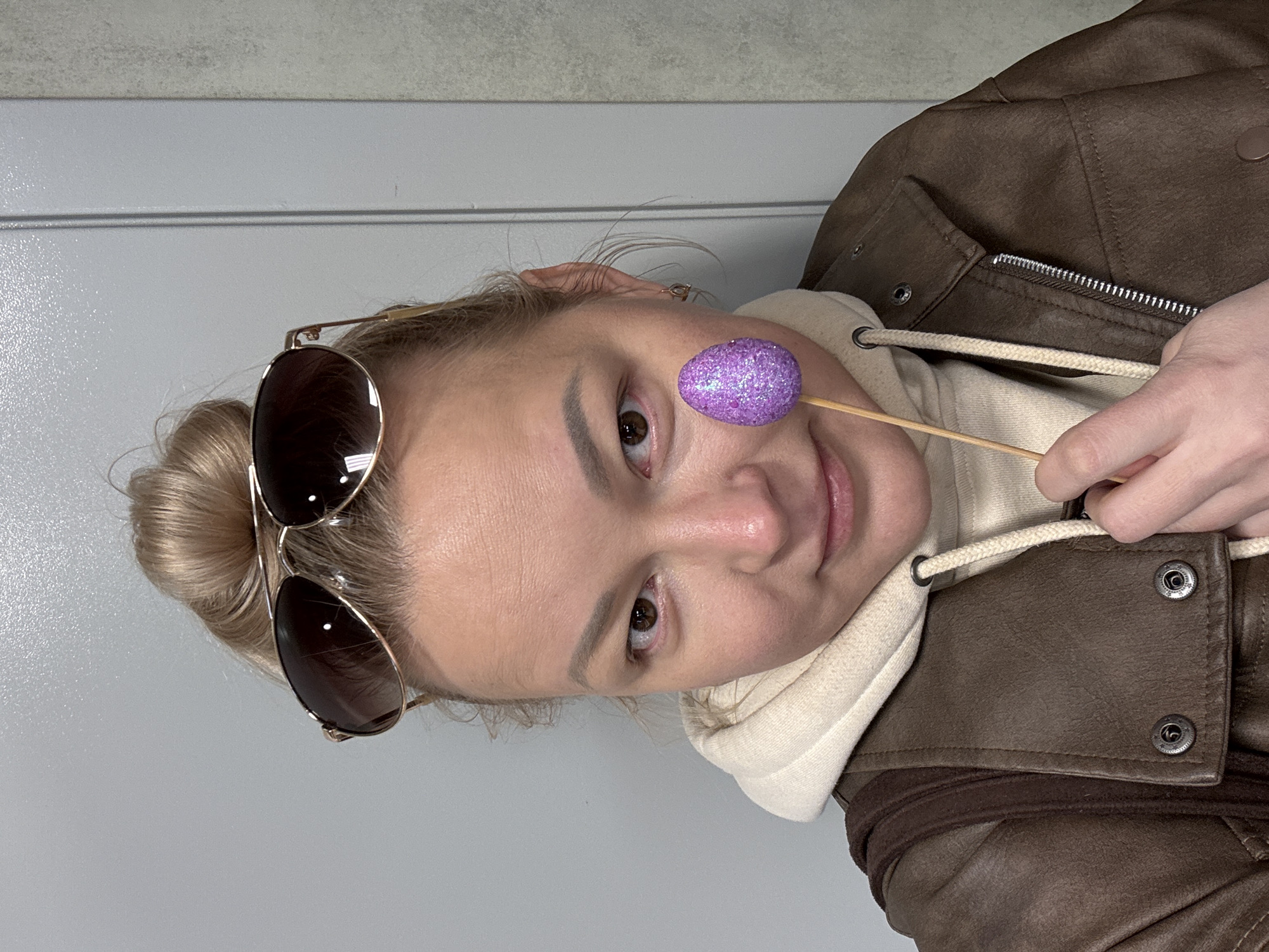 A woman with blonde hair pulled back in a bun is holding a small, sparkly purple Easter egg on a stick up to her right eye. She is wearing aviator sunglasses on top of her head. She is wearing a beige hoodie and a brown leather jacket. The background is a light gray wall. The image is oriented sideways. There is no indication of the city Izhevsk, Russia.