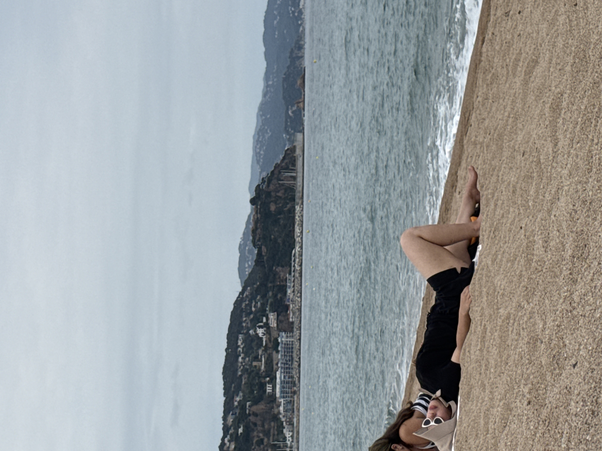Two individuals are lying on a pebble beach. The person in the foreground is wearing a black t-shirt, black shorts, and a light-colored hat with sunglasses. They are lying on their back with their legs bent. The second person, partially visible, is lying on their side wearing a striped bikini top and a dark headscarf. The sea is visible in the background with waves and a shoreline dotted with yellow buoys. In the distance, a hilly coastline with buildings and vegetation is visible, consistent with the topography of Blanes, Spain. The sky is overcast.