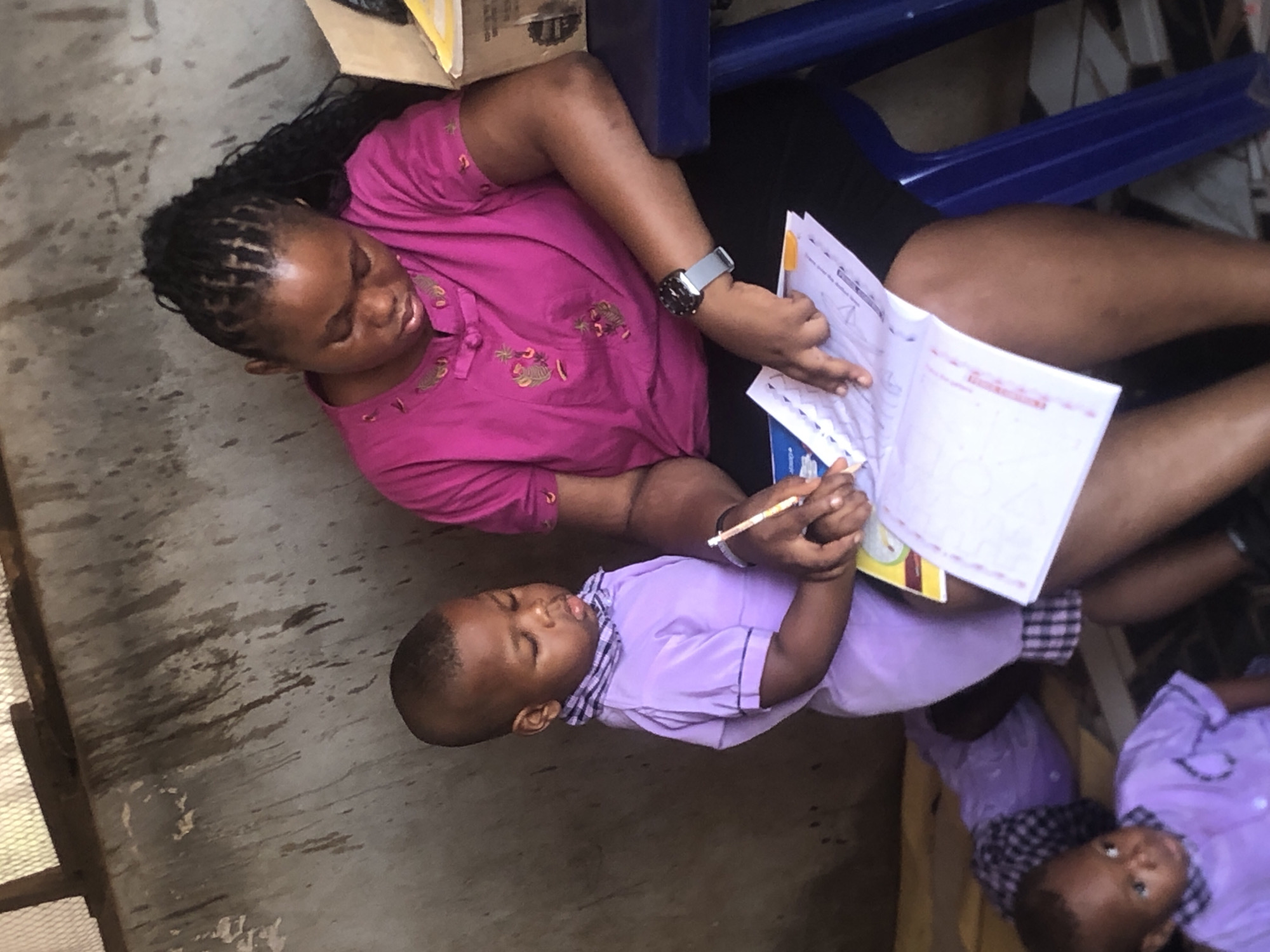 An overhead view captures an adult woman and two young children engaged in a learning activity in an indoor setting, likely in Ukwu-Abwa, Nigeria. The image is slightly rotated clockwise.
The central figure is a woman with dark skin and braided hair, wearing a bright pink short-sleeved top adorned with light embroidery and black shorts or a skirt. She has a silver smartwatch on her left wrist. She is intently looking down at an open textbook or notebook, with her right index finger pointing at content on the left page.
To her left, a young boy with dark skin and short hair is also engrossed in the book. He wears a light purple short-sleeved shirt with a checkered collar and holds a yellow pencil or pen in his right hand, poised as if writing or following along.
Partially visible in the lower right corner is a second, younger child or toddler, also with dark skin and dressed in a light purple top, looking upwards, possibly at the woman.
The activity suggests an educational session, with the woman instructing or assisting the children. The open book displays text and lines, with the word "English" faintly discernible at the top right of the right-hand page. The floor is a worn, grey concrete surface. Elements like a cardboard box and a blue plastic structure (possibly furniture) are visible in the background, hinting at a simple and functional environment. The participants appear focused and engaged in the lesson.