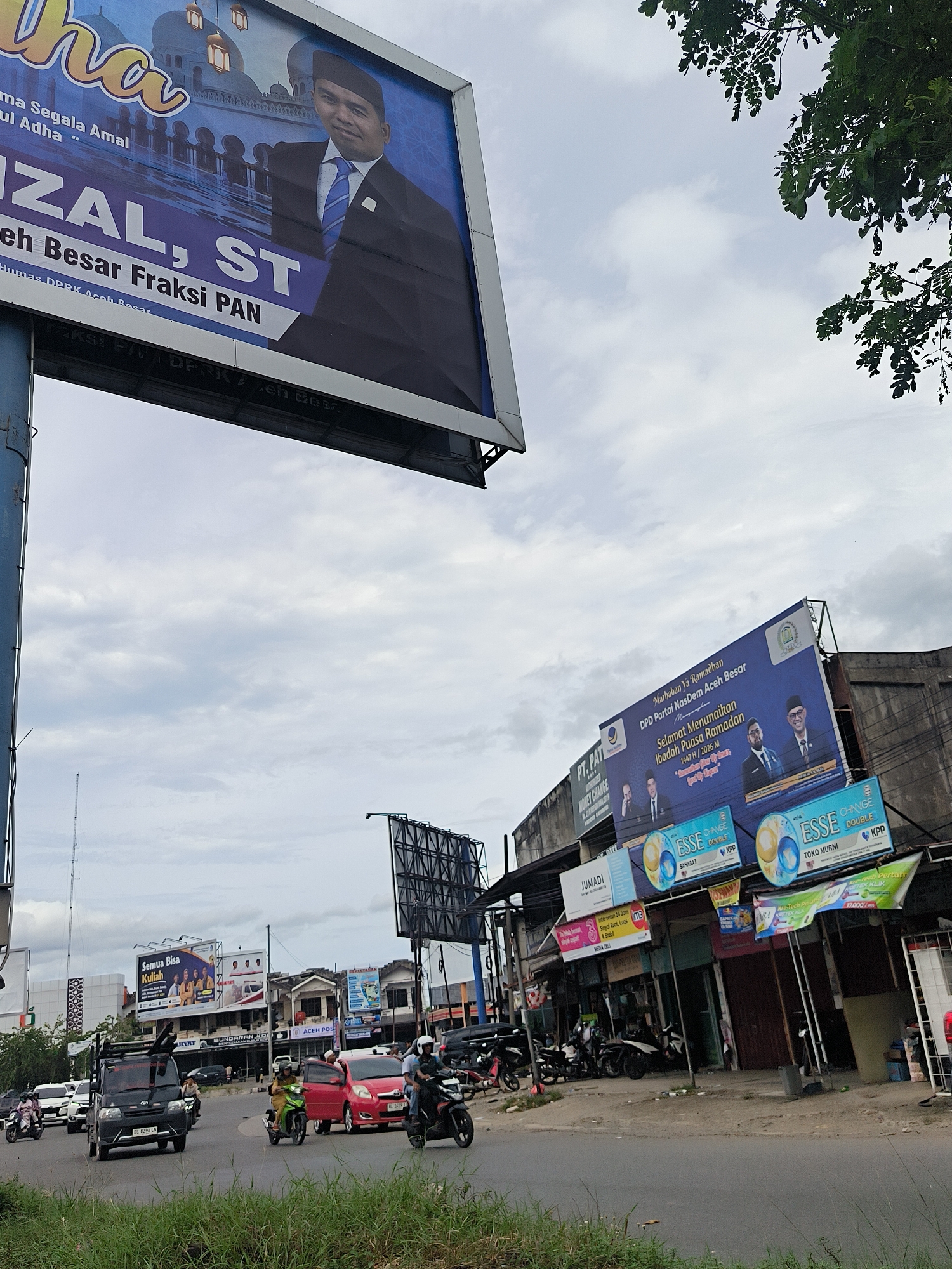 This media file captures a bustling street scene in Lambaro, Indonesia, under an overcast sky. The image shows a road with a grassy median, populated by various vehicles including a black van (license plate BL 8396 LN), a red car, and several motorcycles with riders, indicating active traffic.
Two prominent political billboards dominate the upper half of the image. The large blue billboard on the left features a man identified as "RIZAL, ST" from "Fraksi PAN", with text related to "Idul Adha" and "Humas DPRK Aceh Besar". Another large blue billboard on the right, affixed to a commercial building, displays multiple men and carries the message "Marhaban Ya Ramadhan" and "Selamat Menunaikan Ibadah Puasa Ramadan 1447 H / 2026 M" from "DPD Partai NasDem Aceh Besar".
Below this second billboard, a row of commercial establishments lines the street, featuring various signs. These include "JUMADI", "MEDIA CELL", "MONEY CHANGE", and "ACEH POS". Several advertisements for "ESSE CHANGE DOUBLE" cigarettes are also visible. In the distance, another billboard or sign advertises "Semua Bisa Kuliah".
The sky is cloudy, suggesting a daytime scene without direct sunlight. Tree foliage is visible in the upper right corner, adding a natural element to the urban landscape. The overall scene depicts a busy commercial and transportation hub.