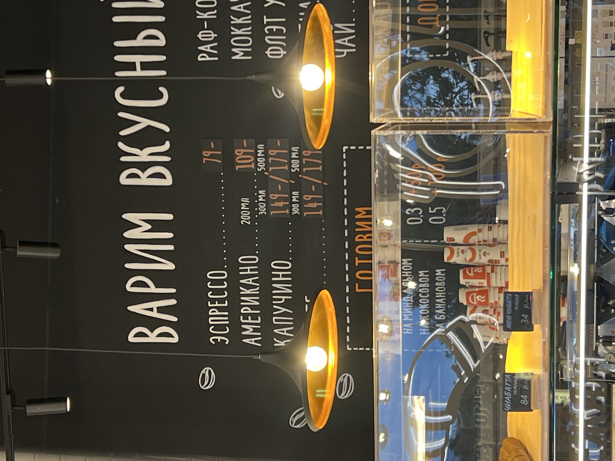 This is a medium shot photograph of a coffee shop's menu board and display case. The menu board is black with white lettering listing coffee drinks like "ЭСПРЕССО," "АМЕРИКАНО," and "КАПУЧИНО," along with prices and sizes. To the right, other coffee drinks like "РАФ-КО" and "МОККА" are listed. Below the main menu, "ГОТОВИМ" is written in orange, indicating food preparation. In the display case below, there are labels for "ЧИАБАТТА" and "МИНИ ЧИАБАТТА" with prices. There are also stacks of coffee cups and reflections of trees and the interior of the shop in the glass. The overall environment suggests a casual, modern cafe. There are no people clearly visible in the frame. The image was taken in Косой Карьер Снт, Russia.