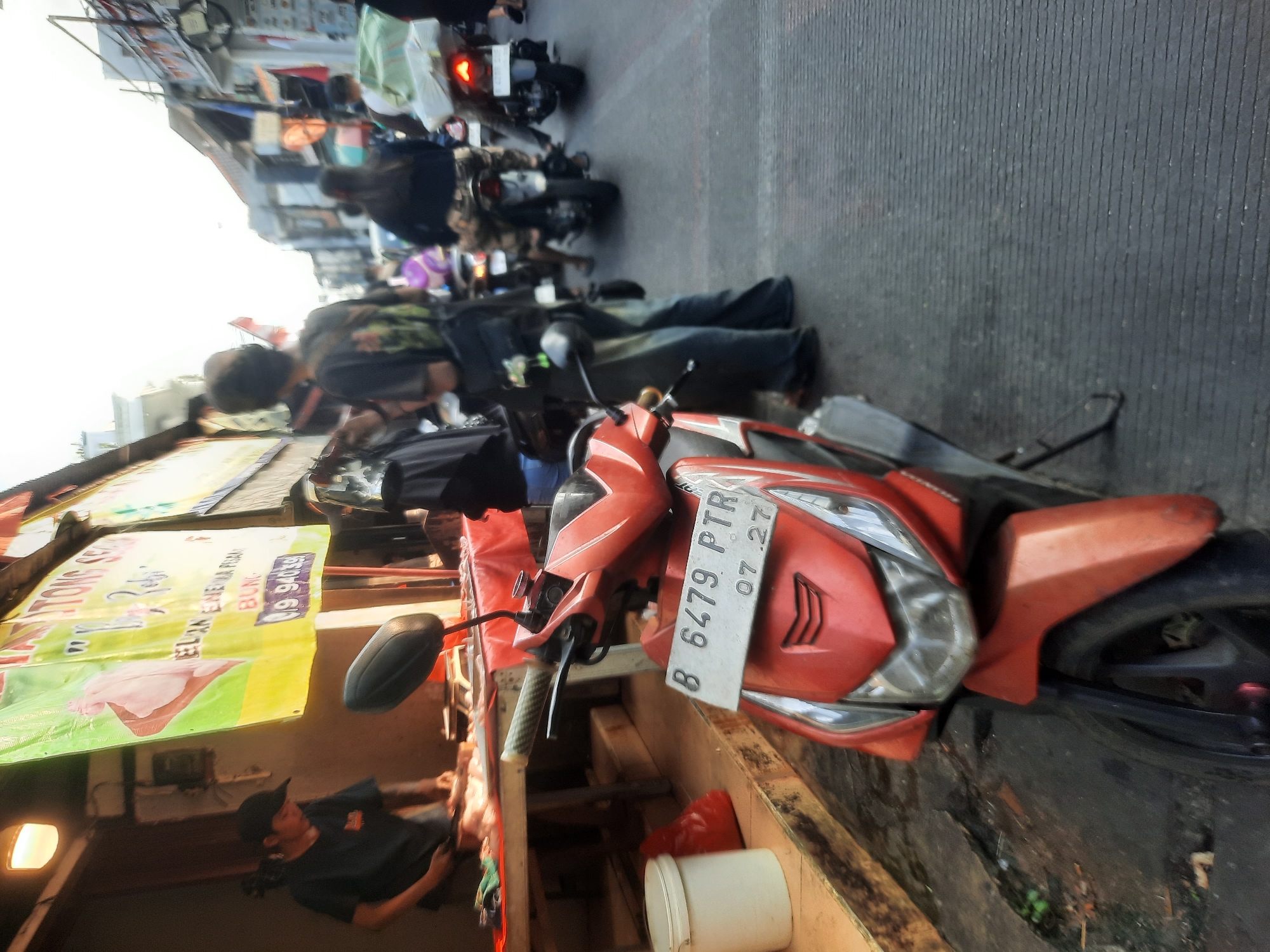 The image captures a street scene in North Jakarta, Indonesia, likely during the daytime given the lighting. In the foreground, a red scooter is parked, its license plate reading "B 6479 PTR 07.27". To the left of the scooter, a person wearing a dark t-shirt and a black cap is working behind a wooden counter, with a red tarp spread over it. Various items, including a white bucket and a red plastic bag, are visible on the ground near the counter.
Behind the counter, a large yellow banner with Indonesian text and an image of a hand holding a piece of meat is displayed. Further down the street, several other motorbikes are present, with people riding them. Some individuals are seen from the back, with one person in the middle ground wearing a dark t-shirt with a graphic design and blue jeans. The background shows buildings lining the street, indicative of a commercial or residential area. The overall impression is a busy urban environment with elements of commerce and daily life.