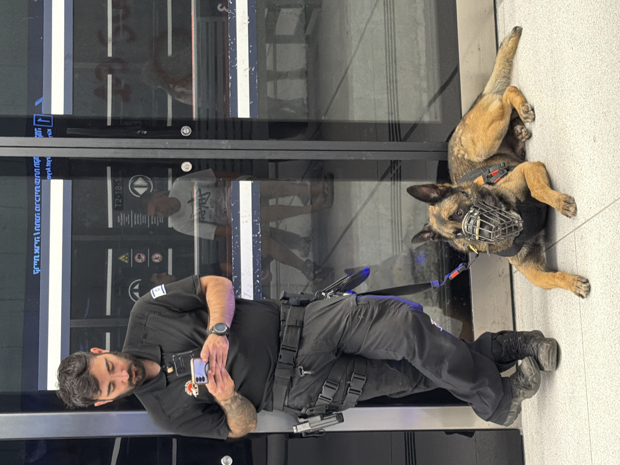 A bearded man wearing a black uniform shirt with an Israeli flag patch, tactical pants, and work boots, stands leaning against a dark reflective surface while looking at his phone. A German Shepherd police dog, wearing a muzzle and a tactical vest, lies on the floor in front of him. The scene appears to be an indoor public transit station, likely in Tel Aviv, Israel, given the signage and the presence of security personnel and a police dog. The man's pants are equipped with a tactical belt holding a holster and other gear. Reflections in the glass doors behind them show other people, suggesting a busy environment.