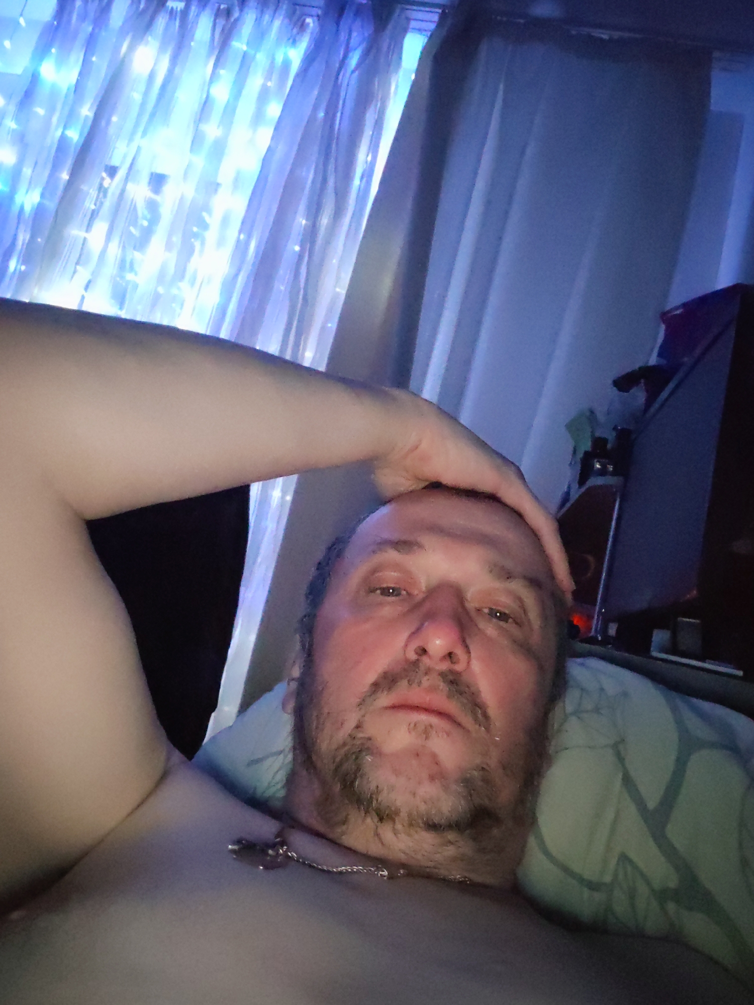 A middle-aged man is lying down, possibly in bed, in what appears to be a dimly lit room. He is shirtless and wearing a silver chain with a pendant. His head rests on a pillow, and his arm is raised, with his hand on his head. Behind him are curtains, one set of which is illuminated with string lights. A desk or nightstand is visible with items on it, including a computer monitor. The location is likely an interior of a residence, possibly in the city of Chaikovsky, Russia, but this cannot be definitively confirmed from the image.