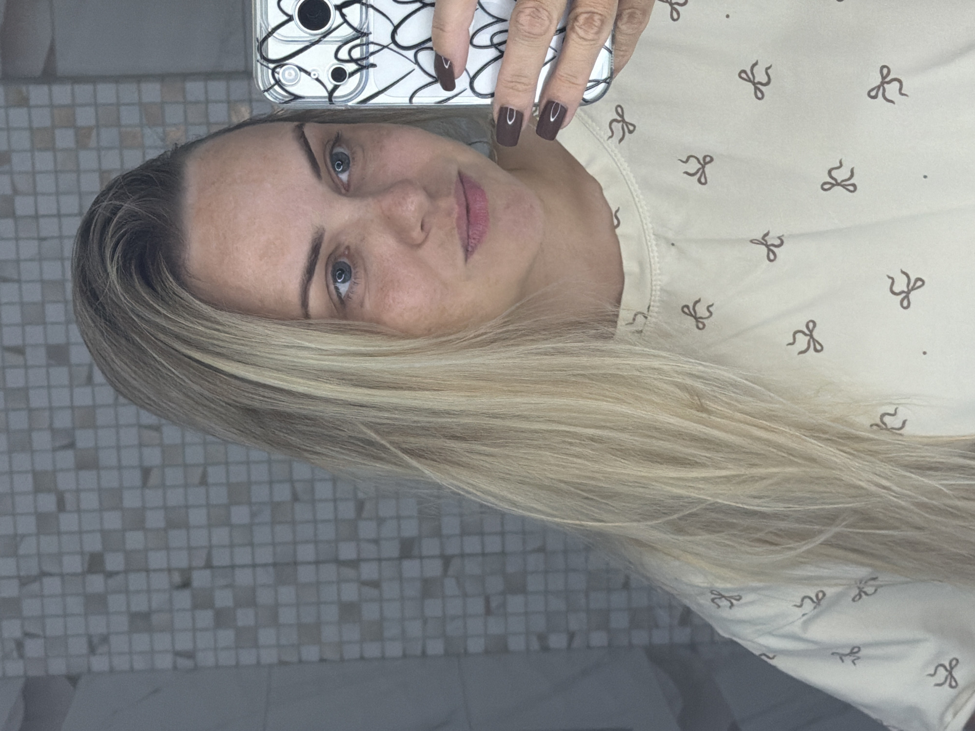 The media file is a selfie taken by a woman in Votkinsk, Russia. She has fair skin with freckles and blue eyes. Her blonde hair is long and styled, framing her face. She is wearing a light-colored shirt with a pattern of dark bows. Her fingernails are painted a dark brown. The background shows a tiled wall, suggesting she is in a bathroom or similar setting. The image appears to be taken indoors, likely during the day. There is no visible text in the media file.