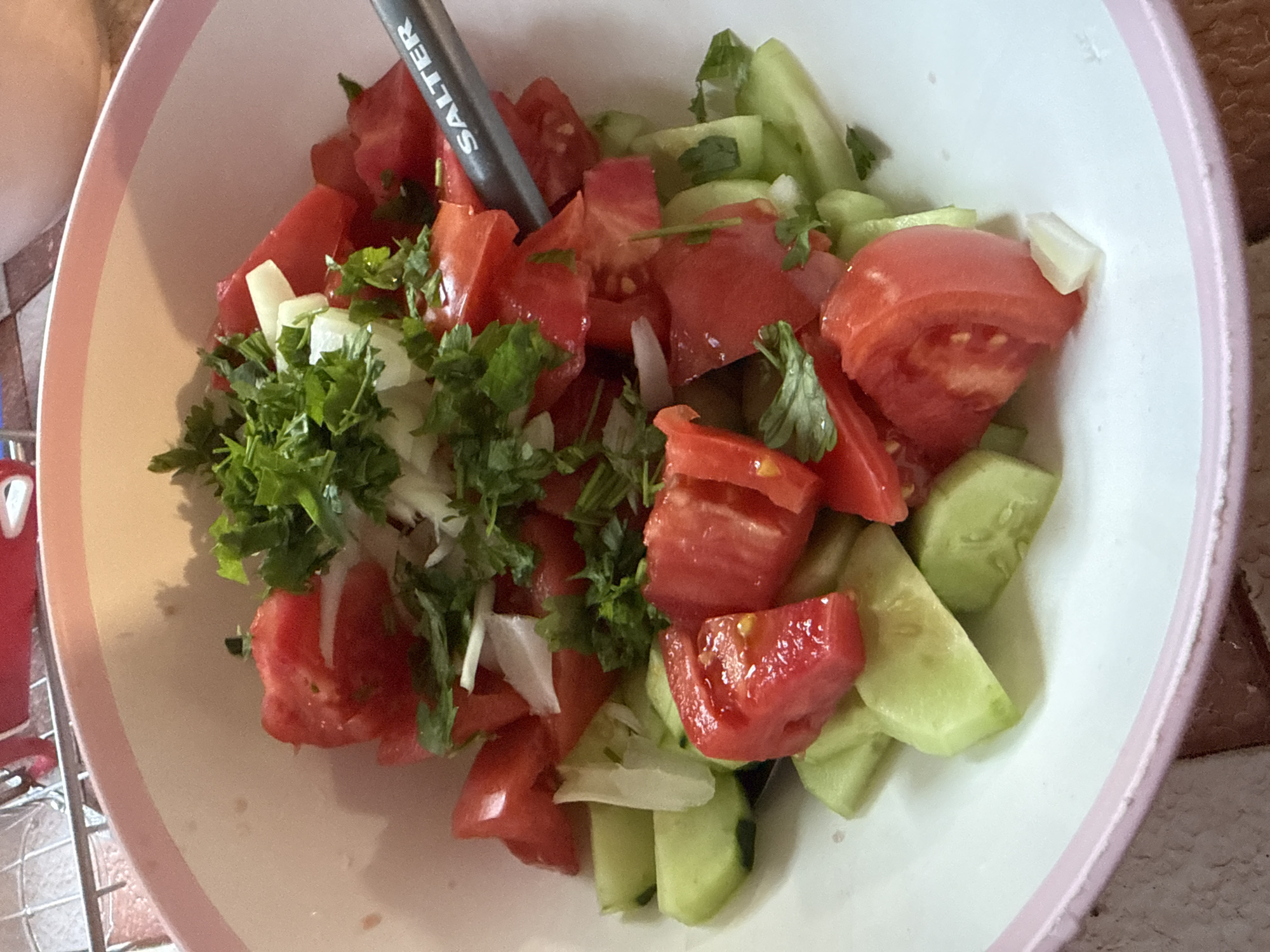 A close-up, overhead shot shows a bowl of chopped salad containing tomatoes and cucumbers, topped with chopped onions and parsley. A grey utensil with the word "SALTER" is partially visible within the bowl, suggesting the salad is being prepared or served. The bowl has a pink rim and is filled with the fresh ingredients. The background is out of focus but appears to be a kitchen counter with some household items. The image does not provide any information about the location in Marinka, Bulgaria.