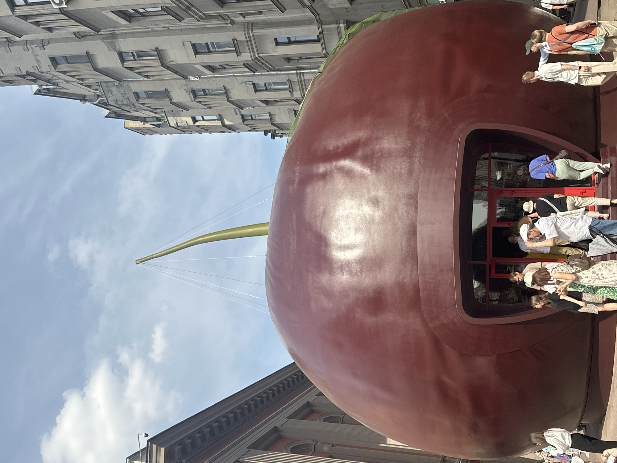 The image presents an outdoor urban scene featuring a massive, glossy, dark red, spherical structure resembling a cherry, positioned centrally. A long, curved, olive-green stem extends upwards from the top of the cherry, supported by white suspension cables. At the base of the cherry, an arched, recessed entrance with red-framed glass doors provides access.
Numerous individuals are observed around the cherry's base and interacting with its entrance. On the left side of the entrance, a woman in a white long-sleeved top and black pants stands with her back to the viewer. Closer to the entrance, a group of casually dressed adults, including a man in a white shirt and beige pants and a woman in a patterned dress, are standing or moving towards the opening. On the right side, several individuals are walking away from the entrance; one man wears a light shirt and beige pants, another a light blue shirt and green cap carrying an orange backpack, and a third an orange t-shirt with a blue bag.
To the left of the cherry, a multi-story building with classical architectural elements, including columns, arched windows, and a prominent cornice, is visible. A security camera is mounted on an upper corner of this building. To the right of the cherry, a tall, multi-story building with a more austere, grayish-beige facade, featuring numerous rectangular windows and slight bay projections, ascends. An air conditioning unit is affixed to an upper section of this building.
The sky above is partly cloudy blue. The ground surrounding the cherry appears to be paved. The scene is illuminated by bright daylight, creating reflections on the cherry's surface. This large cherry installation is located on Bolotnaya Embankment in Moscow, Russia, functioning as a temporary pavilion.