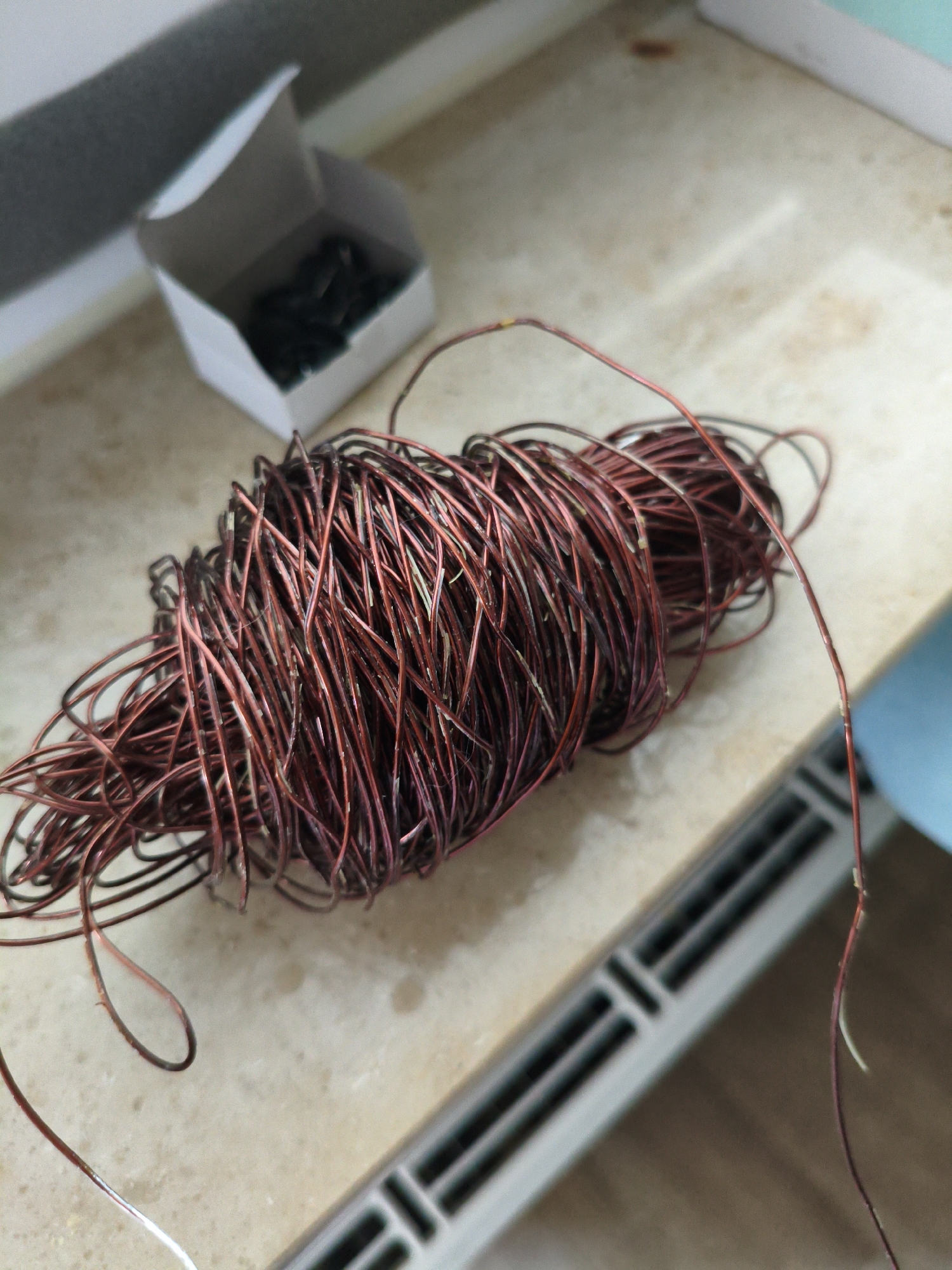 This image captures a close-up view of a tangled ball of thin copper wire, possibly used for electrical work or crafting. The wire is a reddish-brown color, suggesting it might be enameled or aged. Some strands appear to have minor imperfections or debris on them. In the background, a small, open white cardboard box is visible, containing several small, dark objects, the nature of which is unclear. The setting appears to be an indoor surface, possibly a countertop or workbench, with a textured, light-colored material. A subtle shadow falls across the scene from the left. To the right of the wire, a blurred section of a grey and black vent or grate is present. A faint blue object is also visible in the far background. The lighting suggests it is daytime, possibly indoors with ambient light. There is no visible text in the image.
