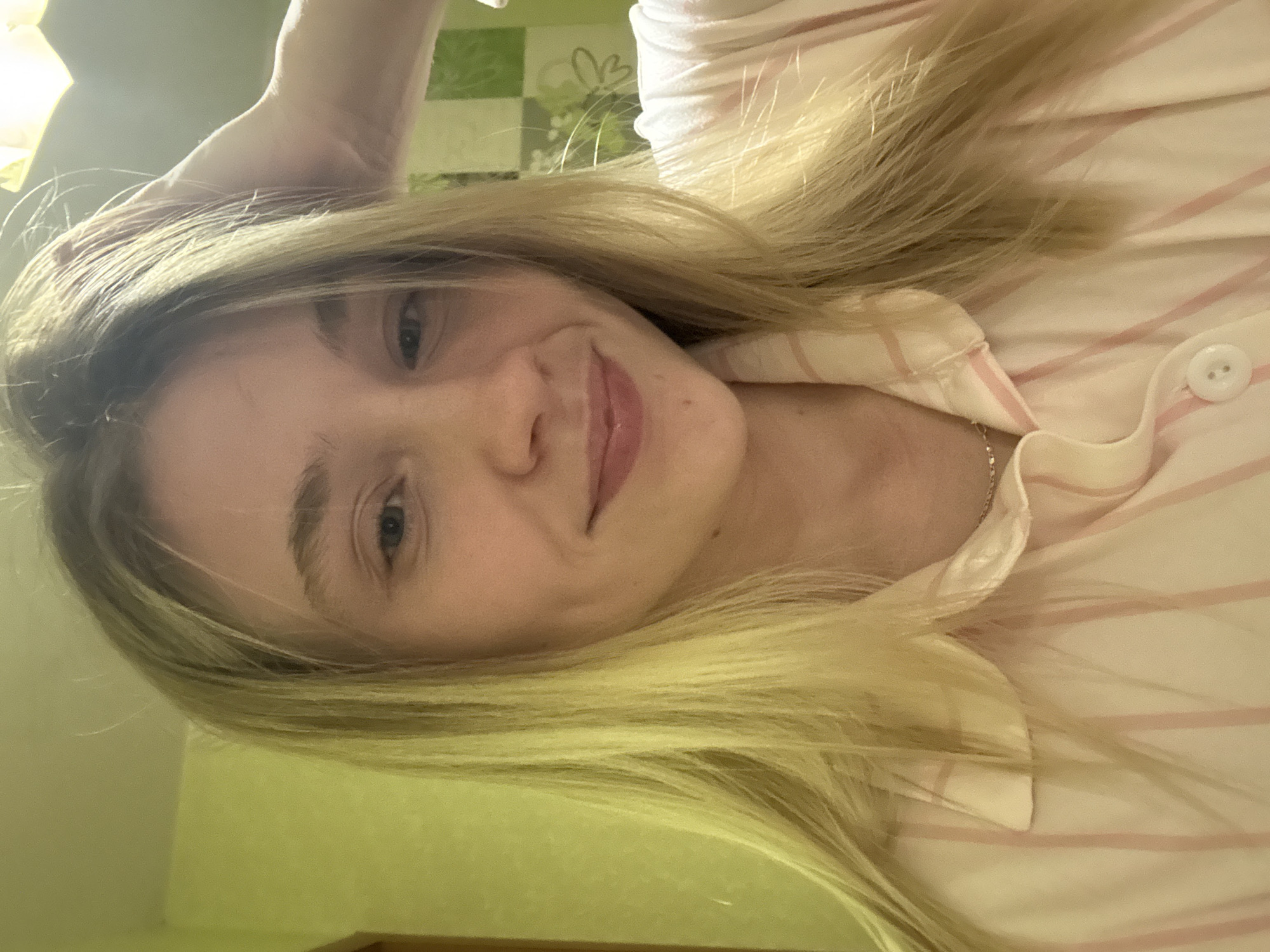 A young woman with long, blonde hair is posing for a selfie in what appears to be an indoor setting. She is wearing a light-colored, striped collared shirt with a button visible at the collar. A delicate necklace is visible at her neck. Her expression is a slight smile. The background features a light green wall and a decorative panel with green and white patterns, including a stylized flower. There is no indication of Izhevsk, Russia in the image.