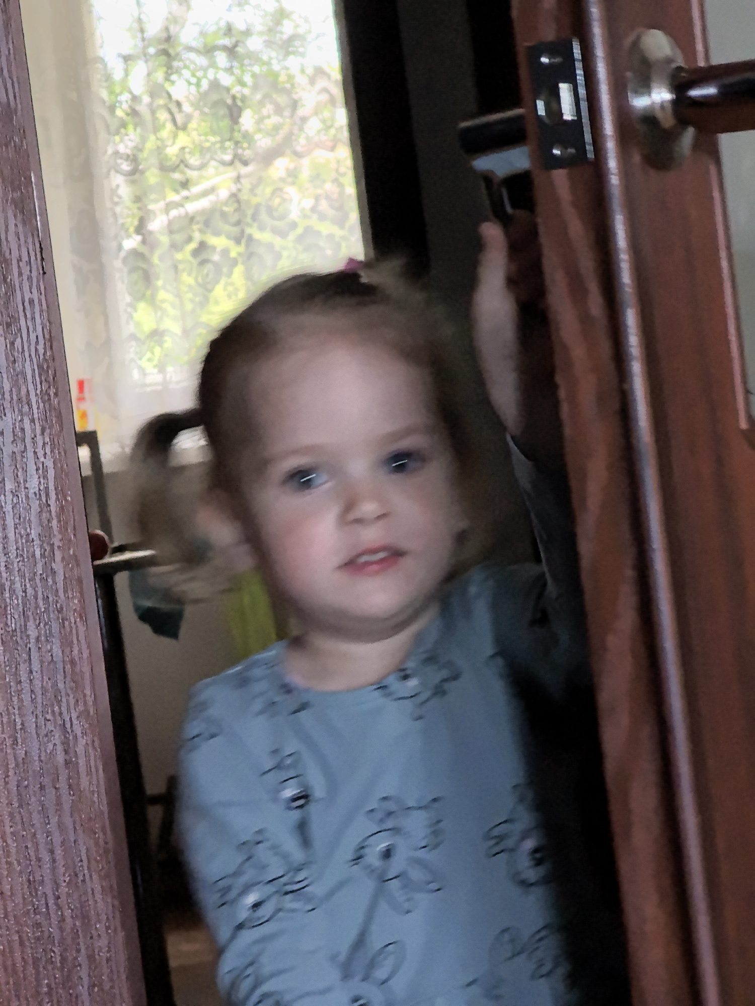 A medium shot shows a young girl with blonde hair in pigtails standing in a doorway. She is wearing a light blue long-sleeved shirt with a black pattern of cartoon animals. The girl is looking towards the right of the frame with a neutral expression. Her right hand is extended slightly. The doorway is partially open, revealing a glimpse of a room with a window covered by a lace curtain. A dark brown doorframe is visible on the right side of the image, with a metallic door handle and lock mechanism. The lighting is soft, suggesting an indoor environment.