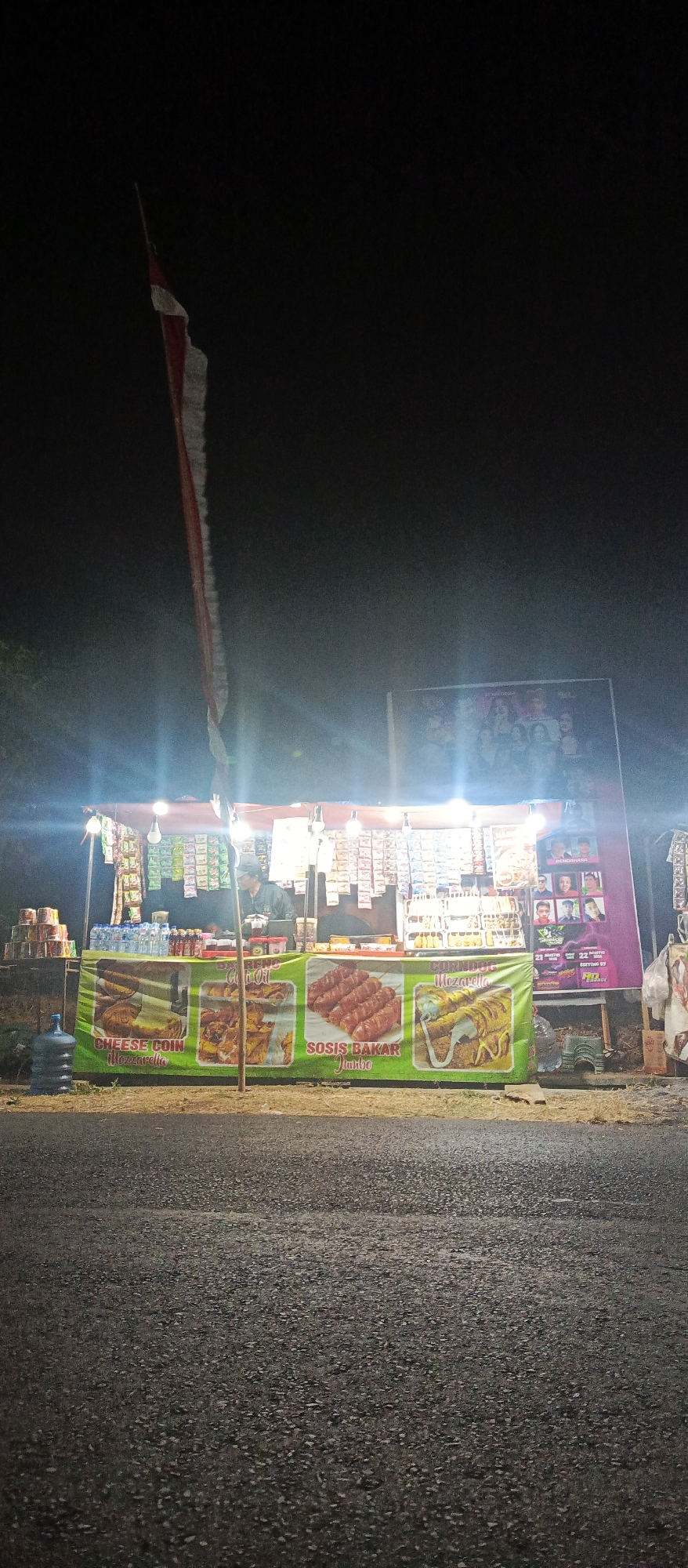 The image depicts a night scene of a food stall situated on the side of a road in Sumbersekar, Indonesia. The stall is illuminated by overhead lights, creating a bright contrast against the dark sky. A red and white Indonesian flag is partially visible, hanging from a pole above the stall.
The stall itself is a long, open-air structure with a covered roof. It is stocked with various food items and beverages. Prominently displayed on the front of the stall are large banners advertising different food products, including "CHEESE COIN Mozzarella," "SOSIS BAKAR Jumbo," and "CORNDOG Mozzarella." Several bottles of water are visible, along with packaged snacks and other products arranged on shelves behind the counter.
A person wearing a cap and a dark jacket is visible behind the counter, appearing to be attending to the stall. The ground in front of the stall is asphalt, suggesting it is located on or near a road. To the right of the stall, a large poster featuring a group of people is displayed. The overall atmosphere suggests a bustling street food environment.