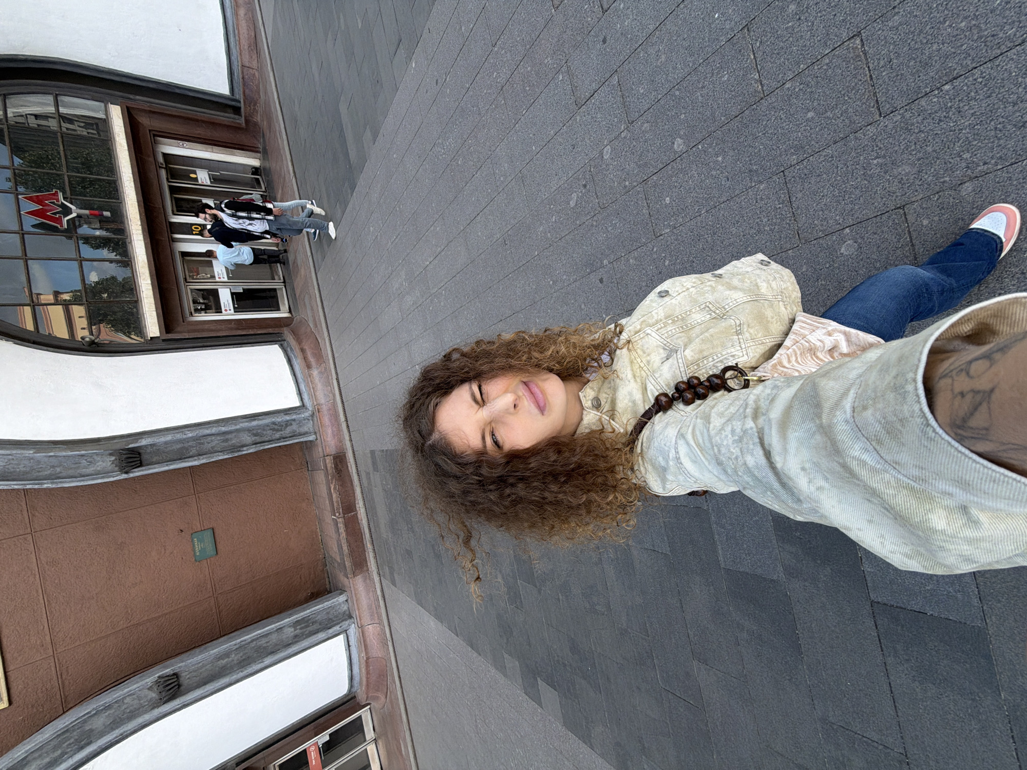 In a selfie taken from a low angle, a woman with curly brown hair and a playful expression stands on a paved walkway. She is wearing a light-colored denim jacket over a patterned top and blue jeans. A large beaded necklace is visible around her neck, and she carries a bag with a distinctive strap. Behind her, an imposing building features a large arched entrance with large windows displaying the distinctive red 'M' logo of the Moscow Metro. Several people are entering and exiting the building in the background. The environment appears to be an urban setting with dark gray paving stones. The building's architecture is characterized by a combination of brown and white facade elements, with curved concrete arches framing the entrance.